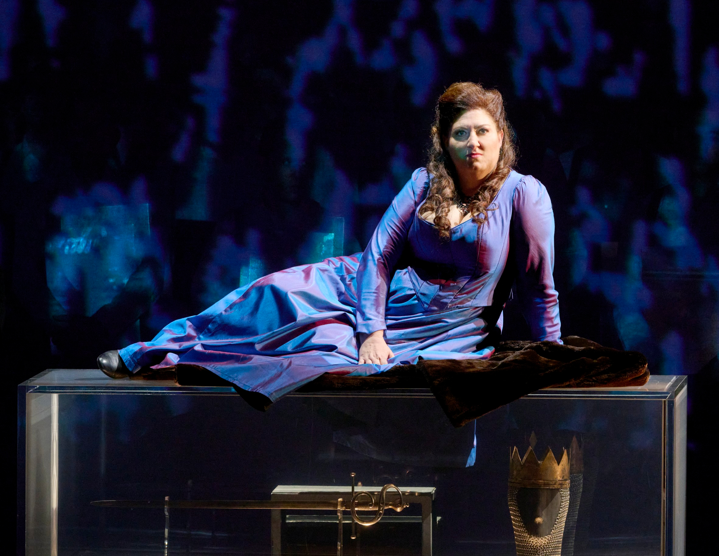 Eine Frau in einem königsblauen Kleid liegt auf einem gläsernen Tisch, unter dem eine goldene Krone sichtbar ist, vor einem dunklen Bühnenhintergrund.