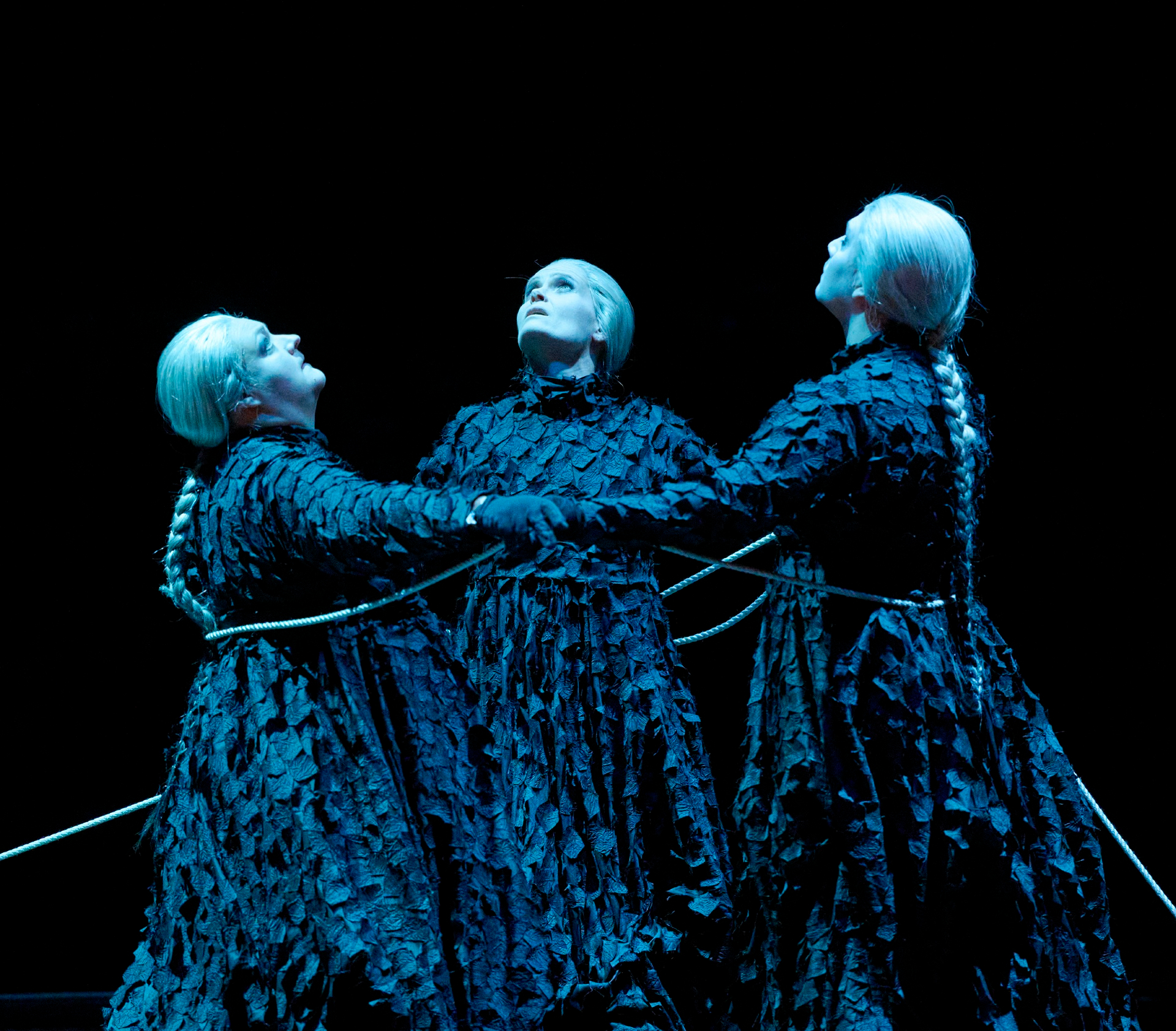 Drei weißhaarige Figuren in dunklen, strukturierten Gewändern stehen auf der Bühne im Kreis, durch ein Seil miteinander verbunden, und blicken dramatisch nach oben.