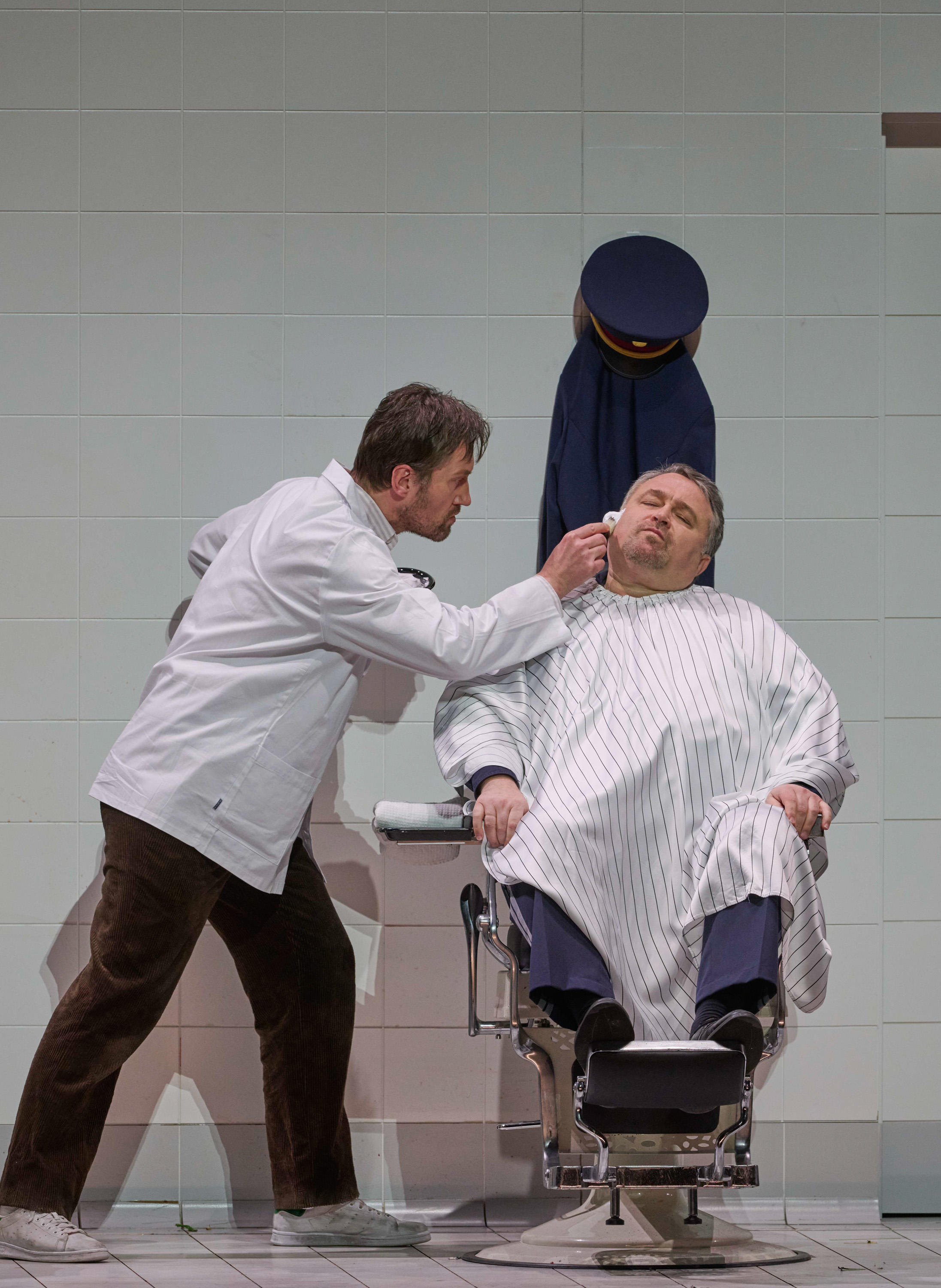 Zwei in weiß gekleidete Männer vor einer gefließten Wand; einer rasiert den Sitzenden im Gesicht.