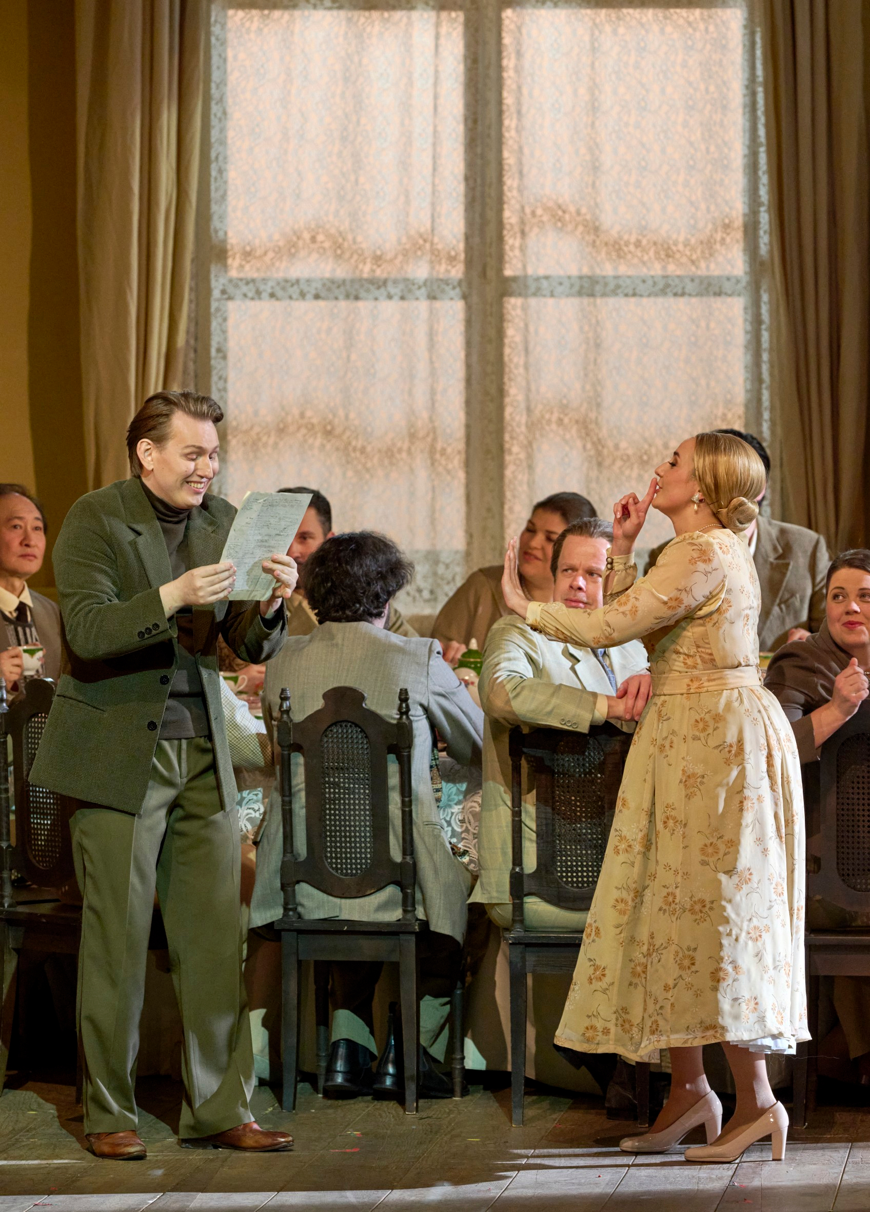 Ein Mann liest mit sichtlicher Freude ein Blatt Papier vor, während eine Frau in hellem Kleid ihn lebhaft gestenreich anspricht; im Hintergrund sitzen mehrere Personen an einem Tisch.