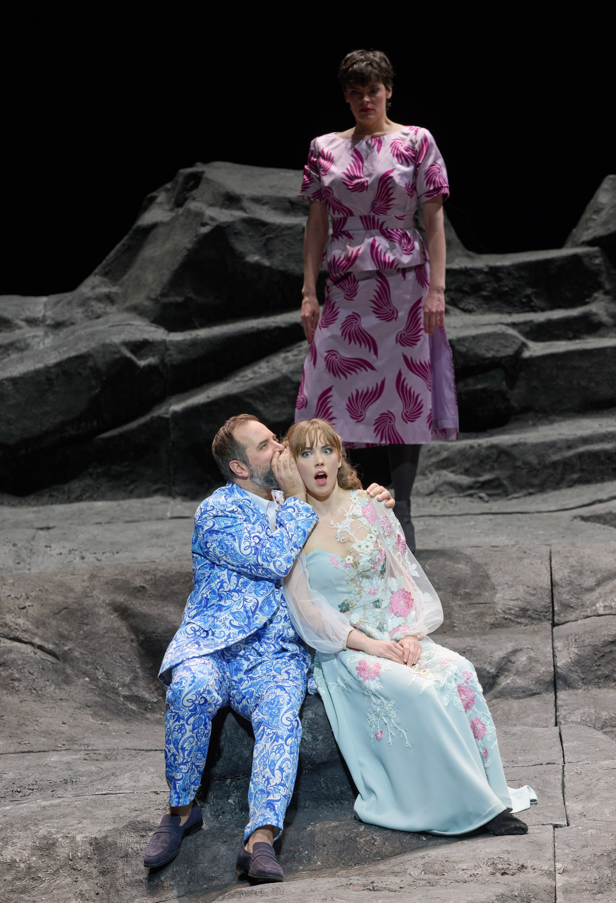 Ein Mann flüstert etwas in das Ohr einer Frau, die überrascht wirkt; im Hintergrund werden sie von einer anderen Frau beobachtet.