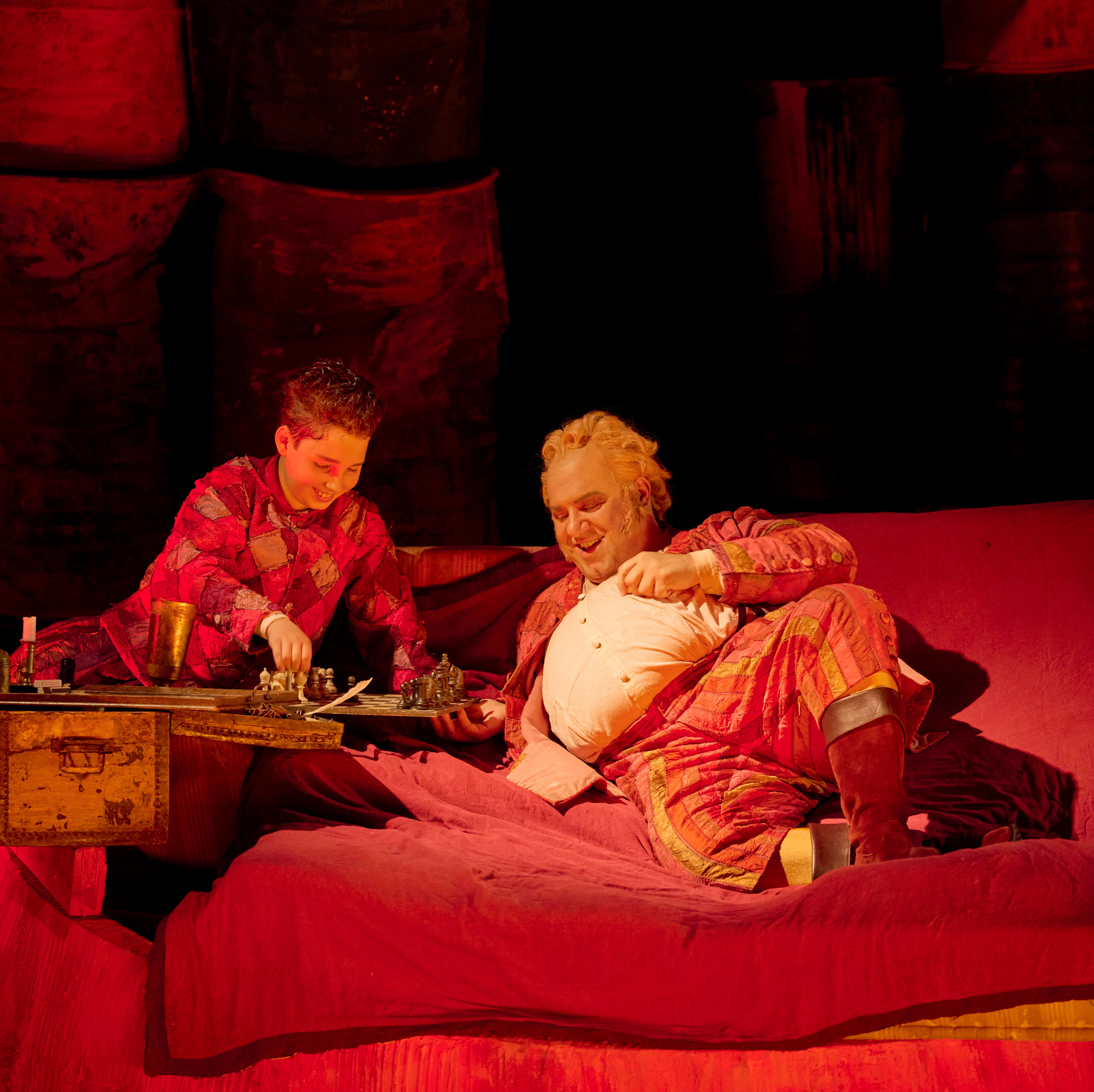 Zwei Personen in farbenfroher Kleidung sitzen entspannt auf einem roten Bett und genießen eine Mahlzeit auf der Bühne.