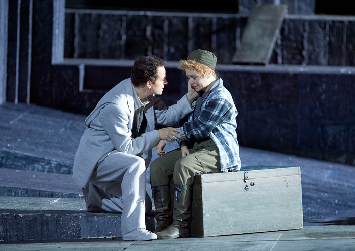 Ein Mann in hellem Anzug kniet vor einem Kind in Holzfällerhemd und Mütze und hält sanft dessen Gesicht.
