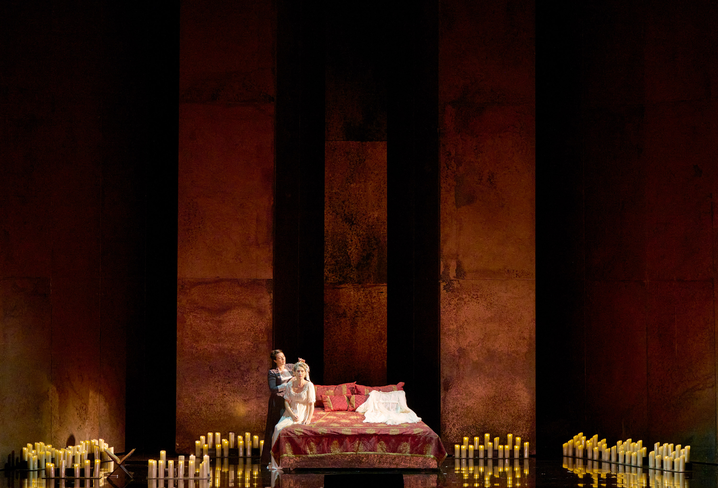 Eine Frau in einem weißen Kleid sitzt auf dem Bett. Hinter ihr steht eine weitere Frau. Der Raum ist gefüllt mit brennenden Kerzen.