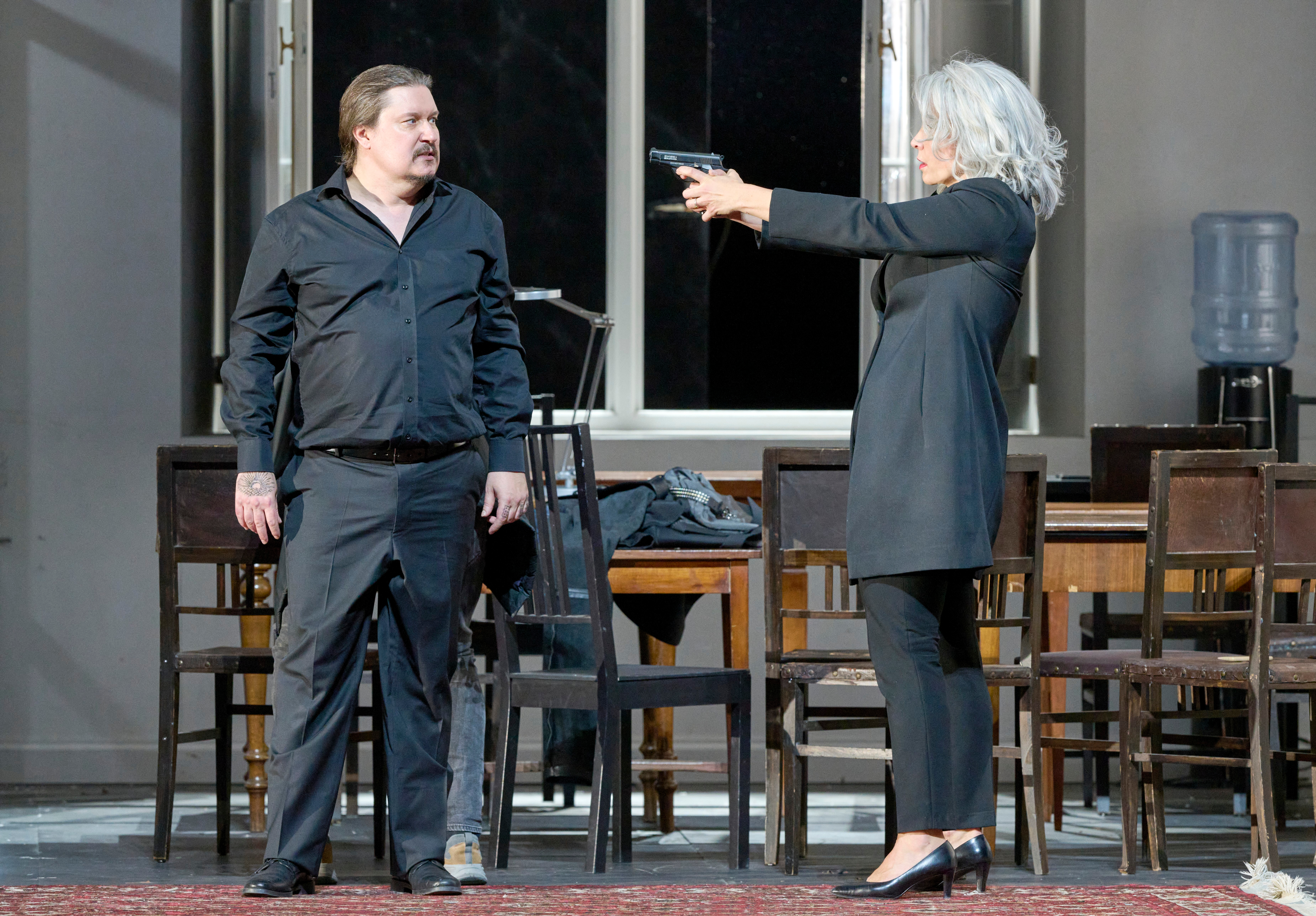 Eine Frau mit silbergrauem Haar richtet eine Pistole auf einen Mann in schwarzer Kleidung, der ihr ruhig gegenübersteht.