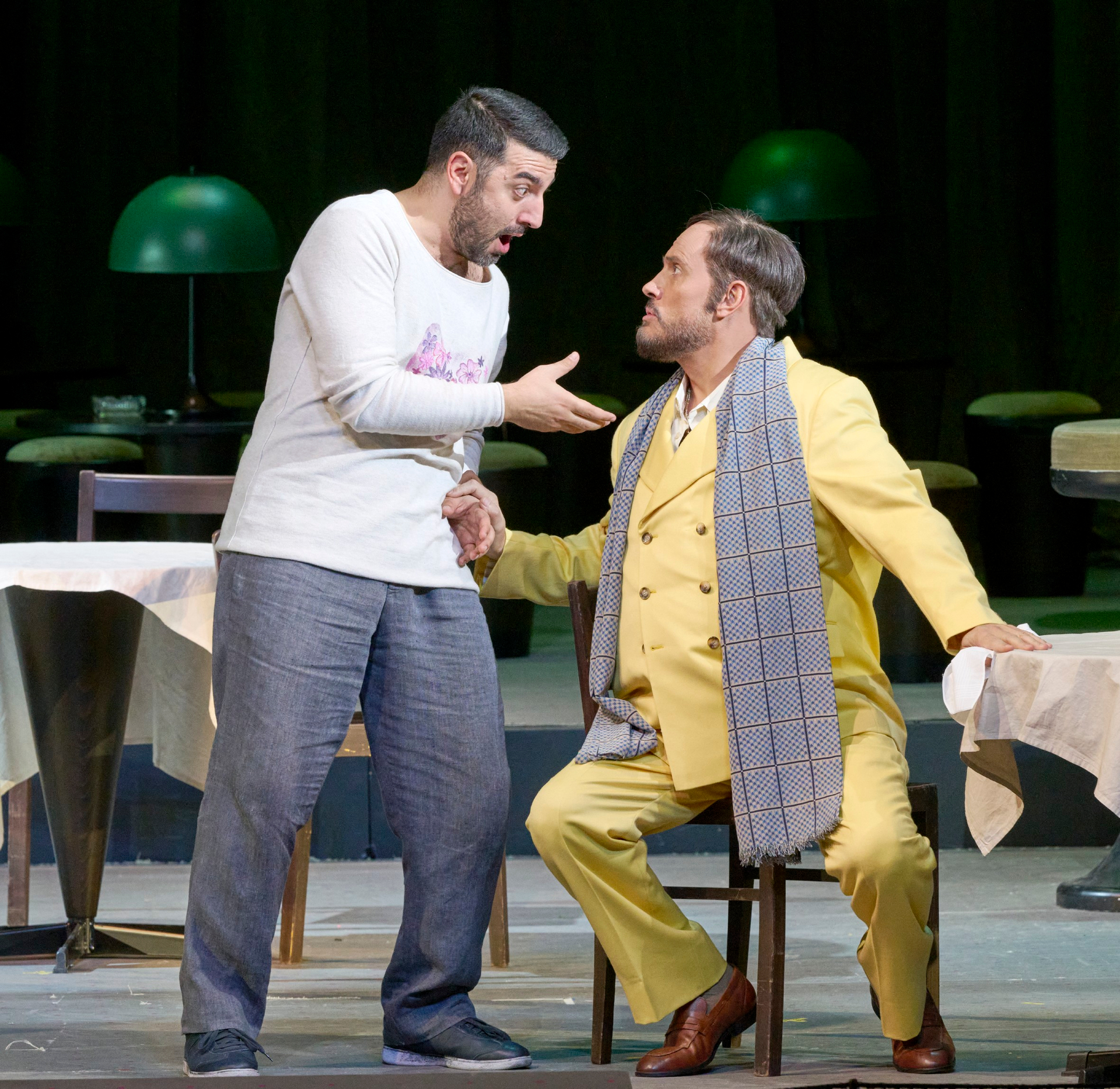 Ein stehender Mann spricht zu einem anderen Mann im gelben Anzug, der auf einem Holzstuhl sitzt.