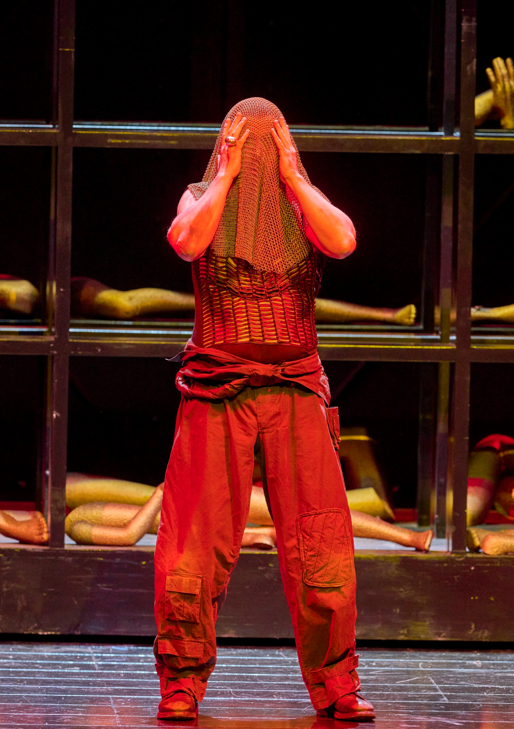 Eine Person in rotem Licht bedeckt ihr Gesicht mit einem Tuch, während im Hintergrund Gliedmaßen hinter Gittern hervorragen.