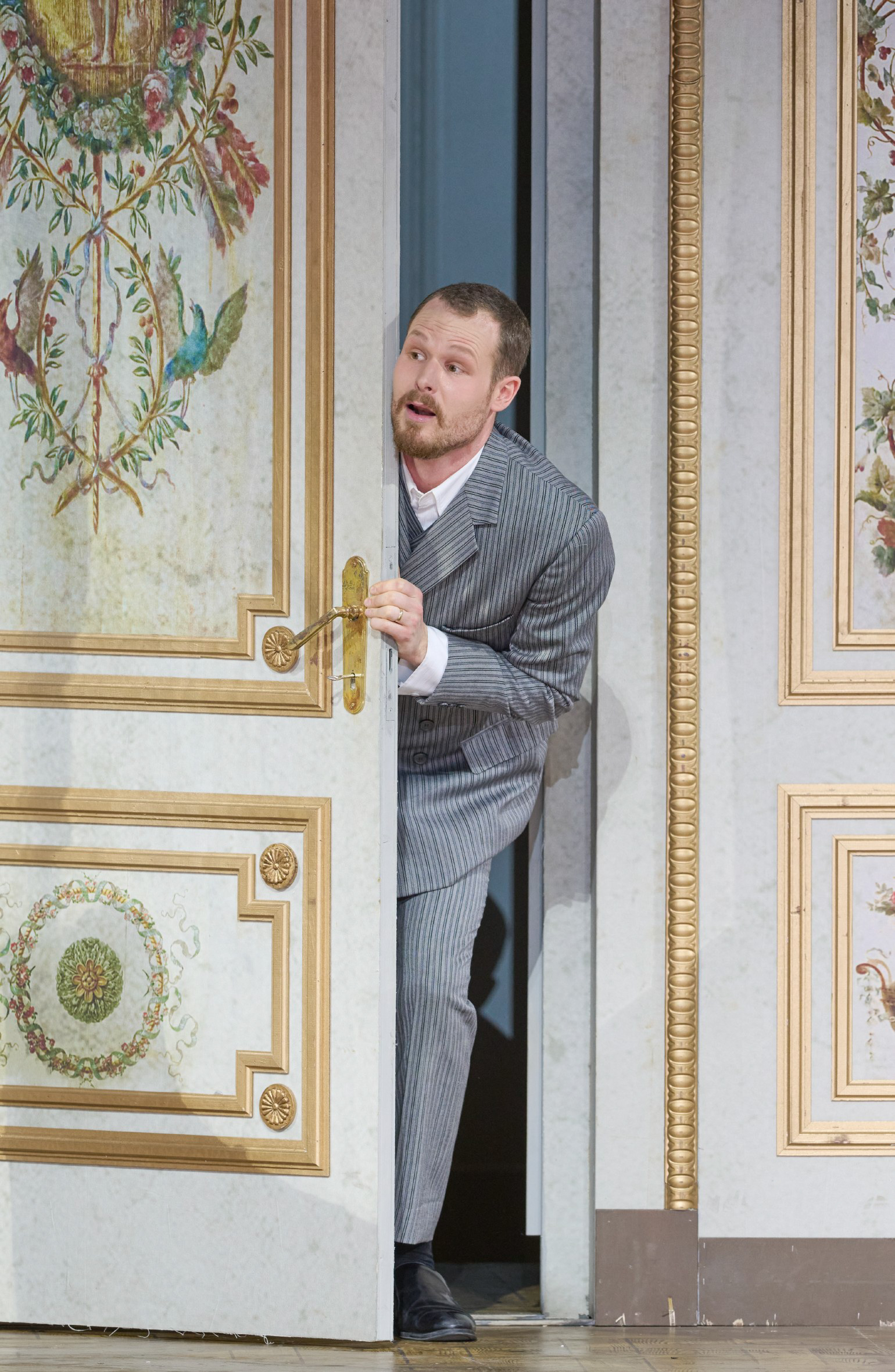 Ein Mann im grauen Anzug lugt neugierig durch eine halb geöffnete, prunkvoll verzierte Tür in einem klassisch dekorierten Raum.