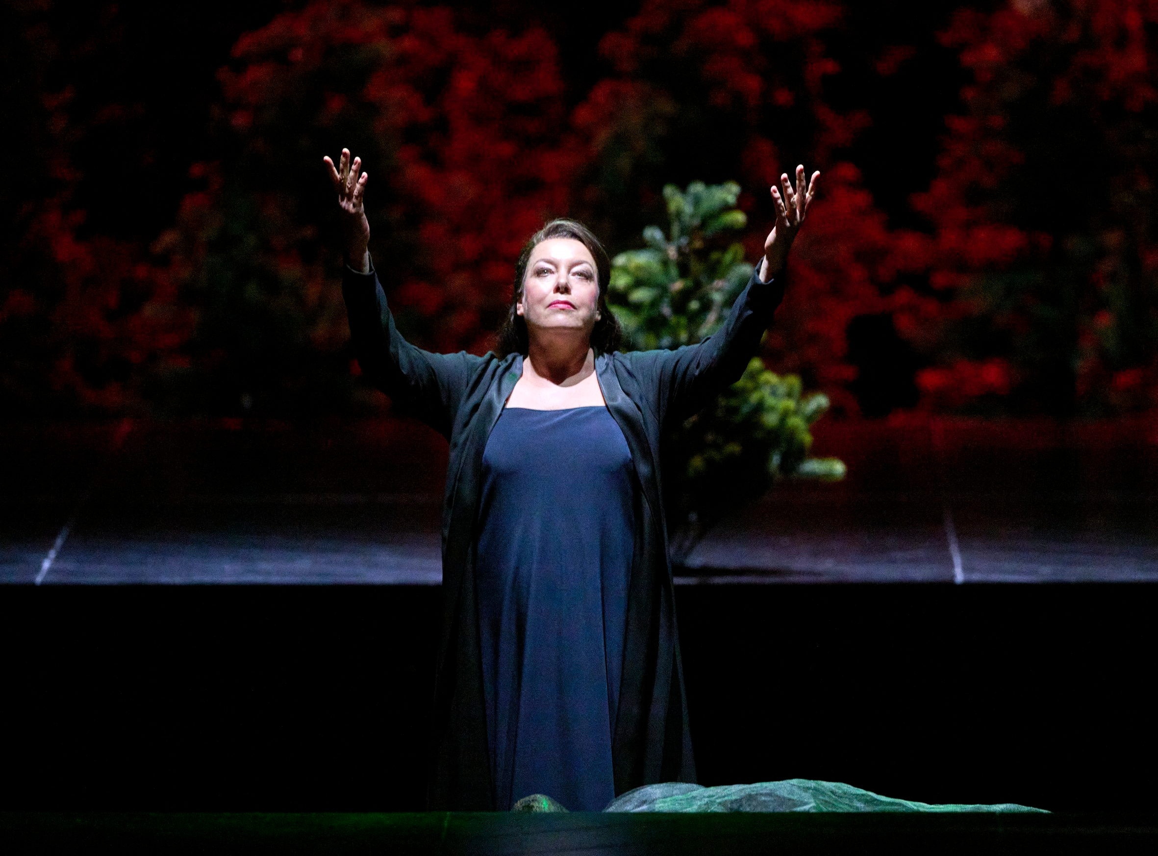 Eine Frau steht auf der Bühne mit erhobenen Armen vor einem Hintergrund aus rotem Laub und wirkt ausdrucksstark und ergriffen.