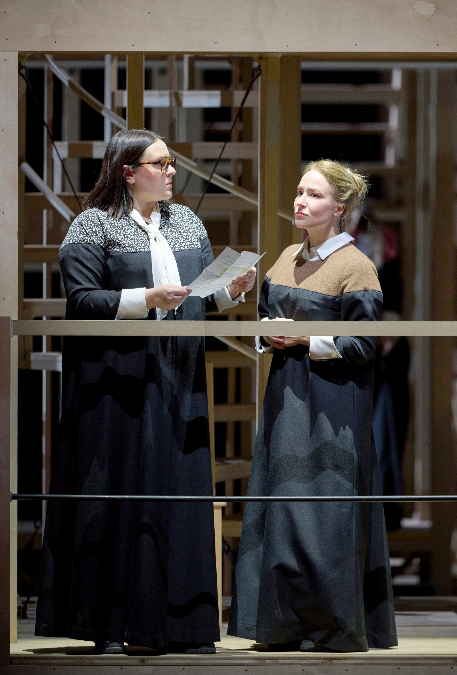 Zwei Frauen in dunkler Kleidung stehen an einem Geländer; eine liest aus einem Blatt Papier, während die andere nachdenklich zuhört.