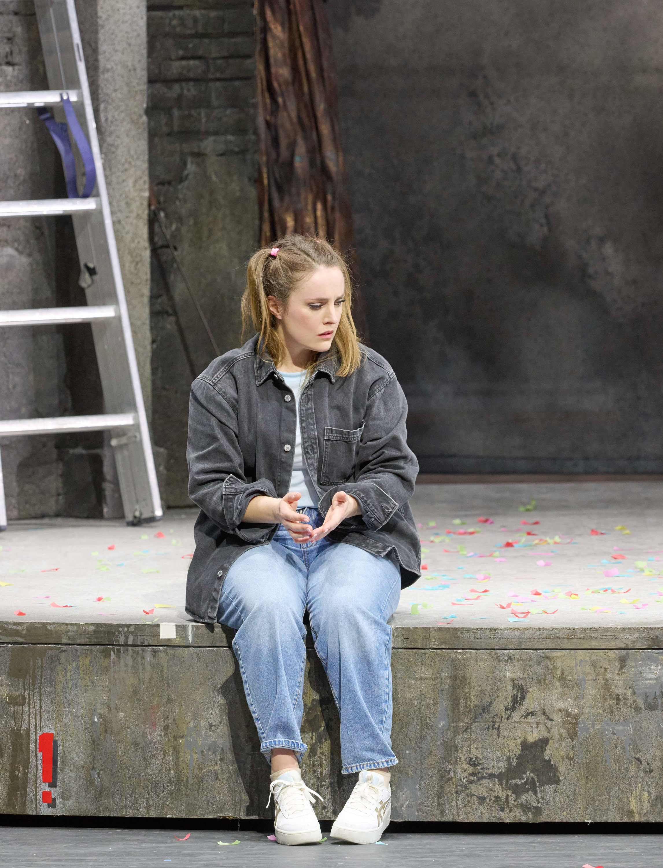 Eine junge Frau mit hellbraunen Haaren sitzt nachdenklich auf einem Podest vor einer grauen Wand, eine Leiter lehnt daneben.