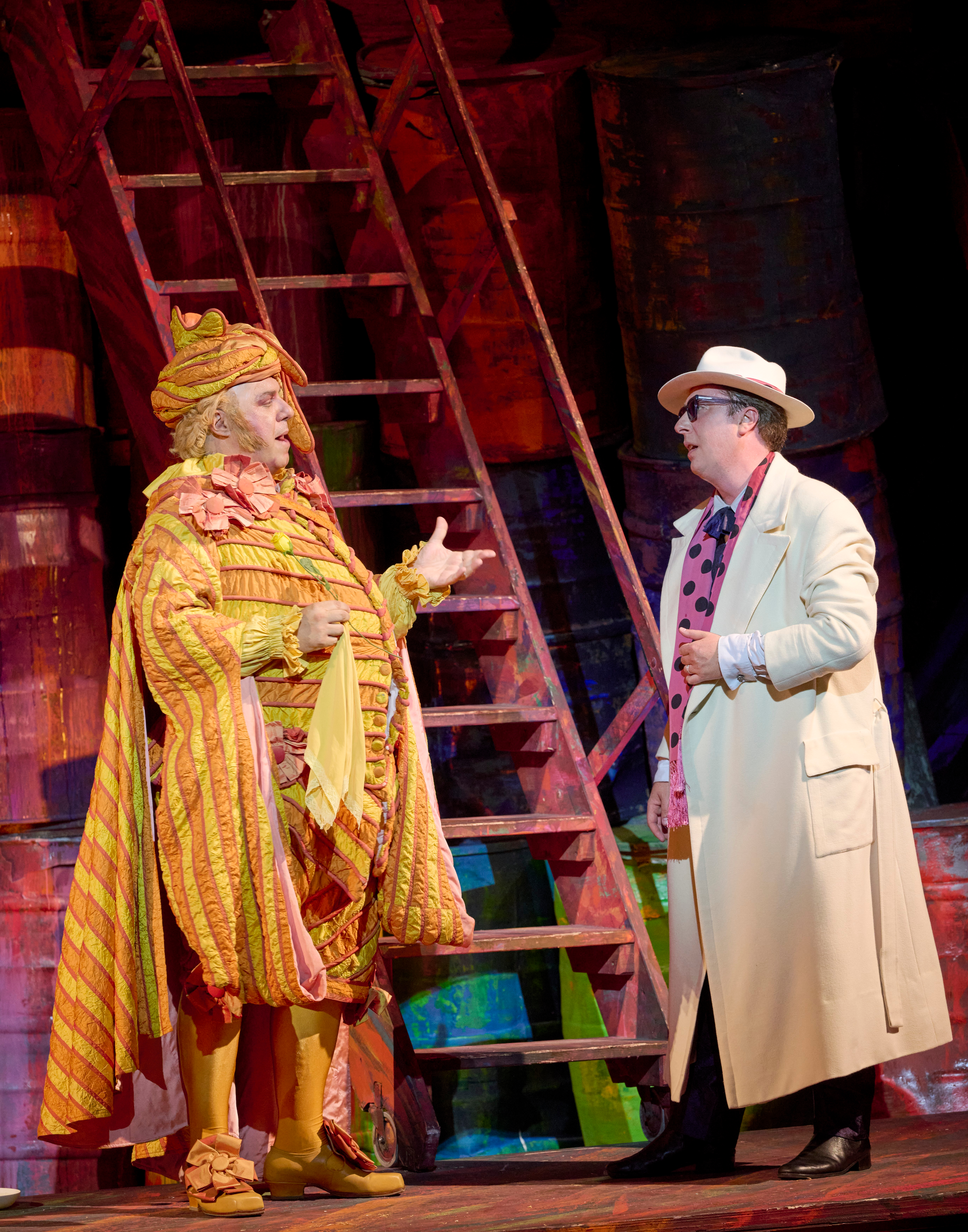 Zwei Darsteller stehen vor einer Holzleiter auf der Bühne; einer trägt ein prachtvolles gelbes Kostüm, der andere einen hellen Mantel und Hut.
