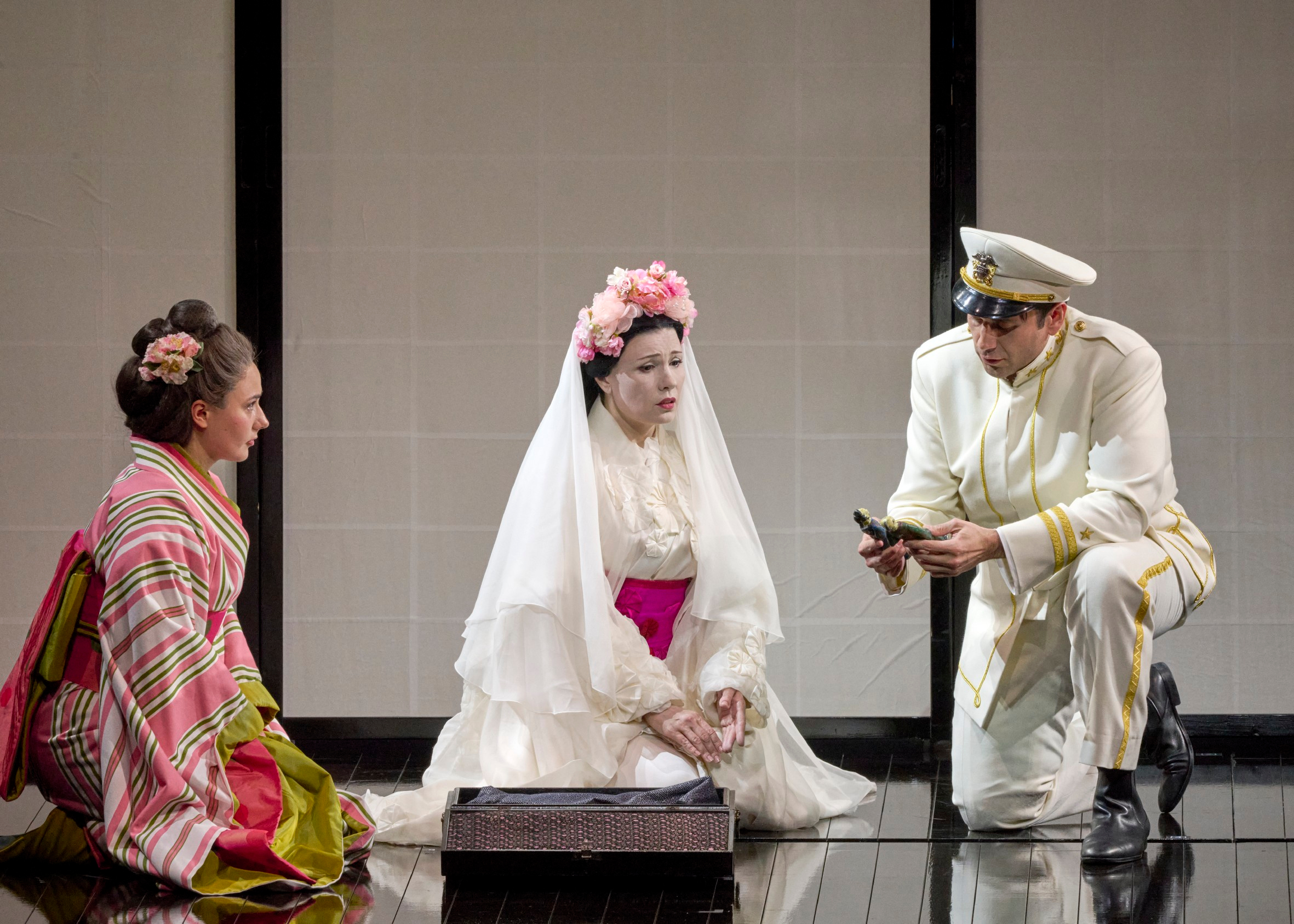 Eine weiß geschminkte Frau im Brautkleid sitzt zwischen einer Frau im Kimono und einem Mann in weißer Uniform auf einer hellen Bühnenfläche.