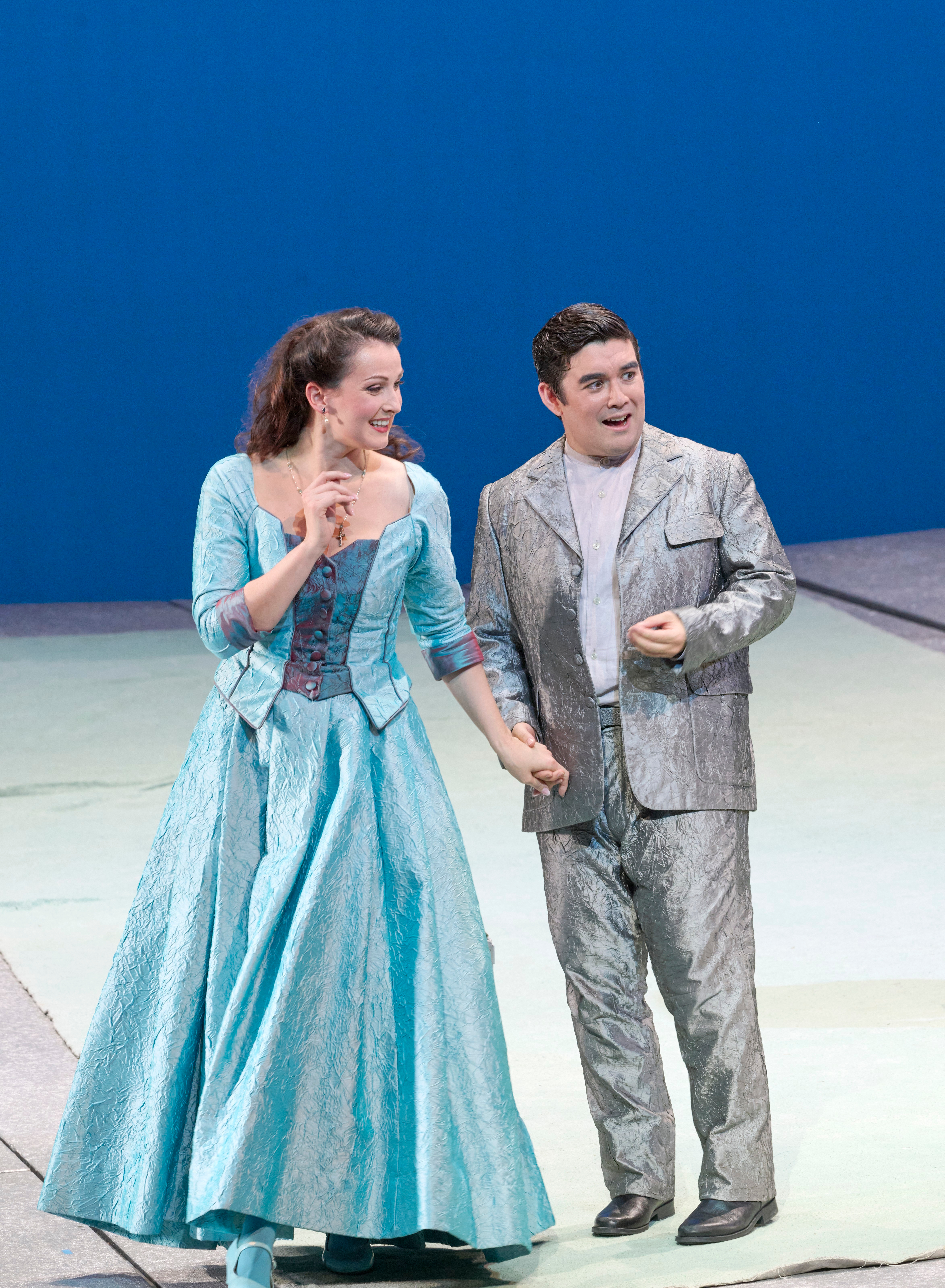 Eine Frau in türkisfarbenem Kleid und ein Mann in silbernem Anzug gehen lächelnd nebeneinander über die Bühne.