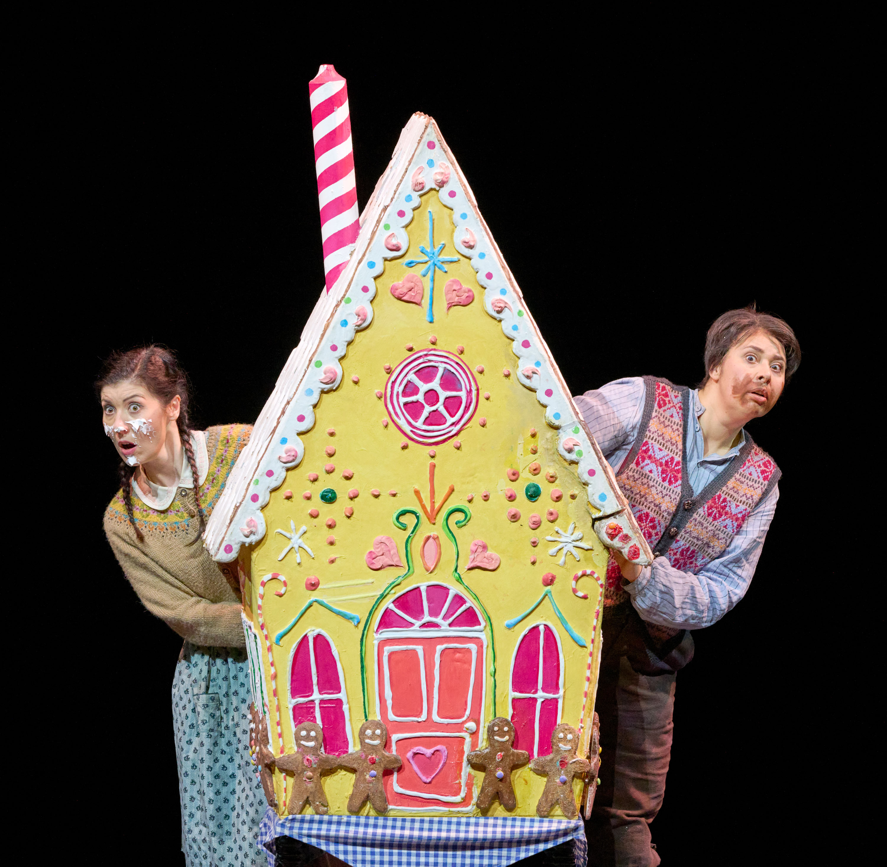 Zwei Kinder blicken neugierig hinter einem bunten, märchenhaften Lebkuchenhaus hervor, das auf der Bühne steht.