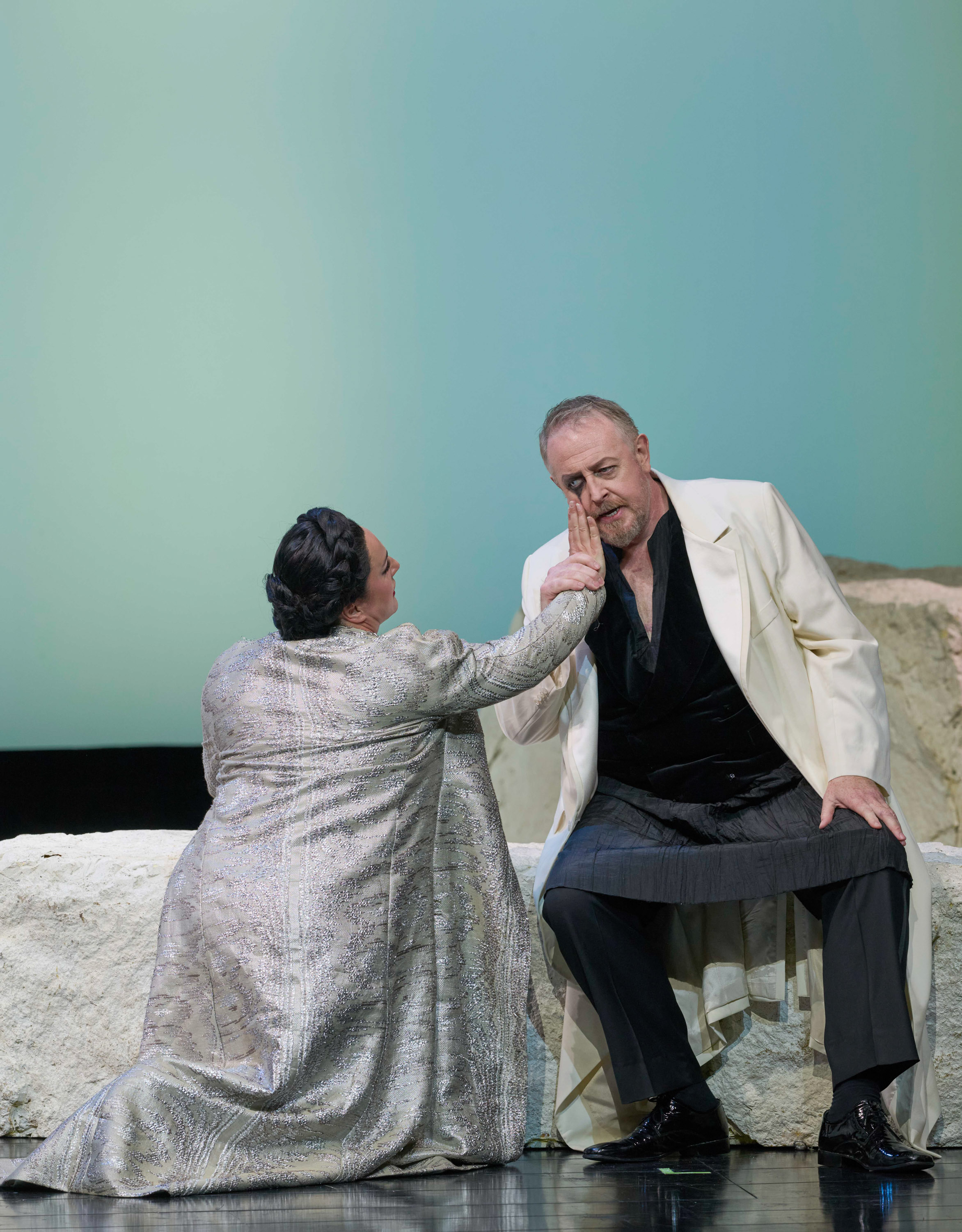 Eine Frau in einem silbernen Mantel berührt zärtlich das Gesicht eines Mannes, der auf einem Felsen sitzt.