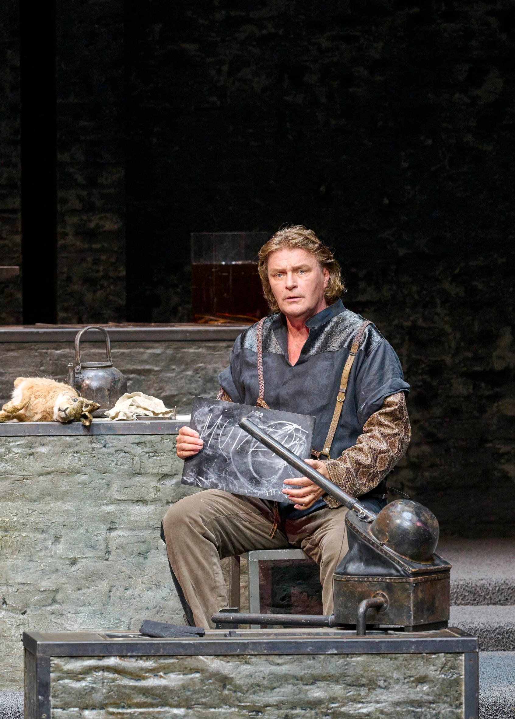 Ein Mann in mittelalterlich wirkender Kleidung sitzt auf einer steinernen Bank und hält eine gravierte Metallplatte mit ernstem Blick.