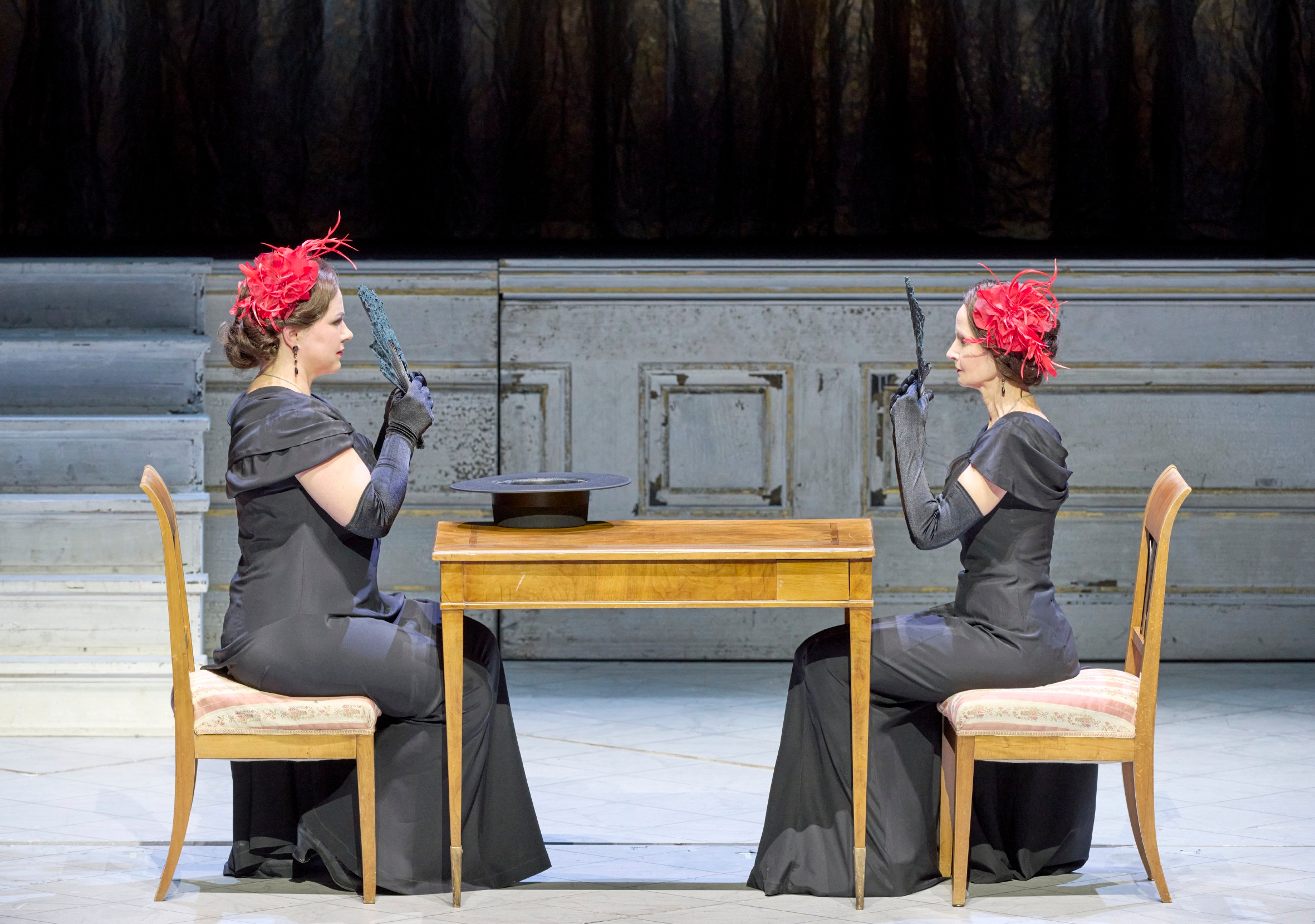 Zwei elegant gekleidete Frauen in Schwarz sitzen sich an einem Holztisch gegenüber.