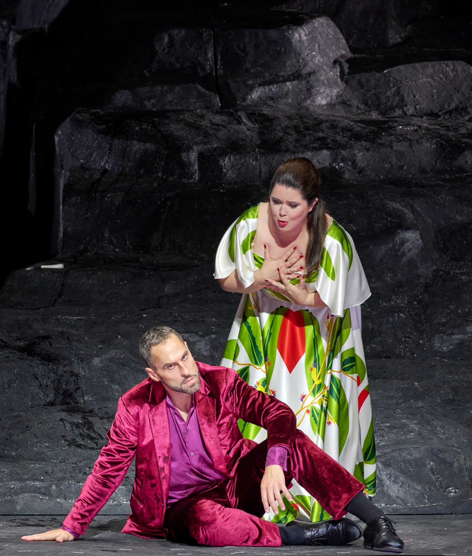 Auf dem Boden sitzt ein Mann im purpurnen Samt-Anzug; über ihn beugt sich eine Frau.
