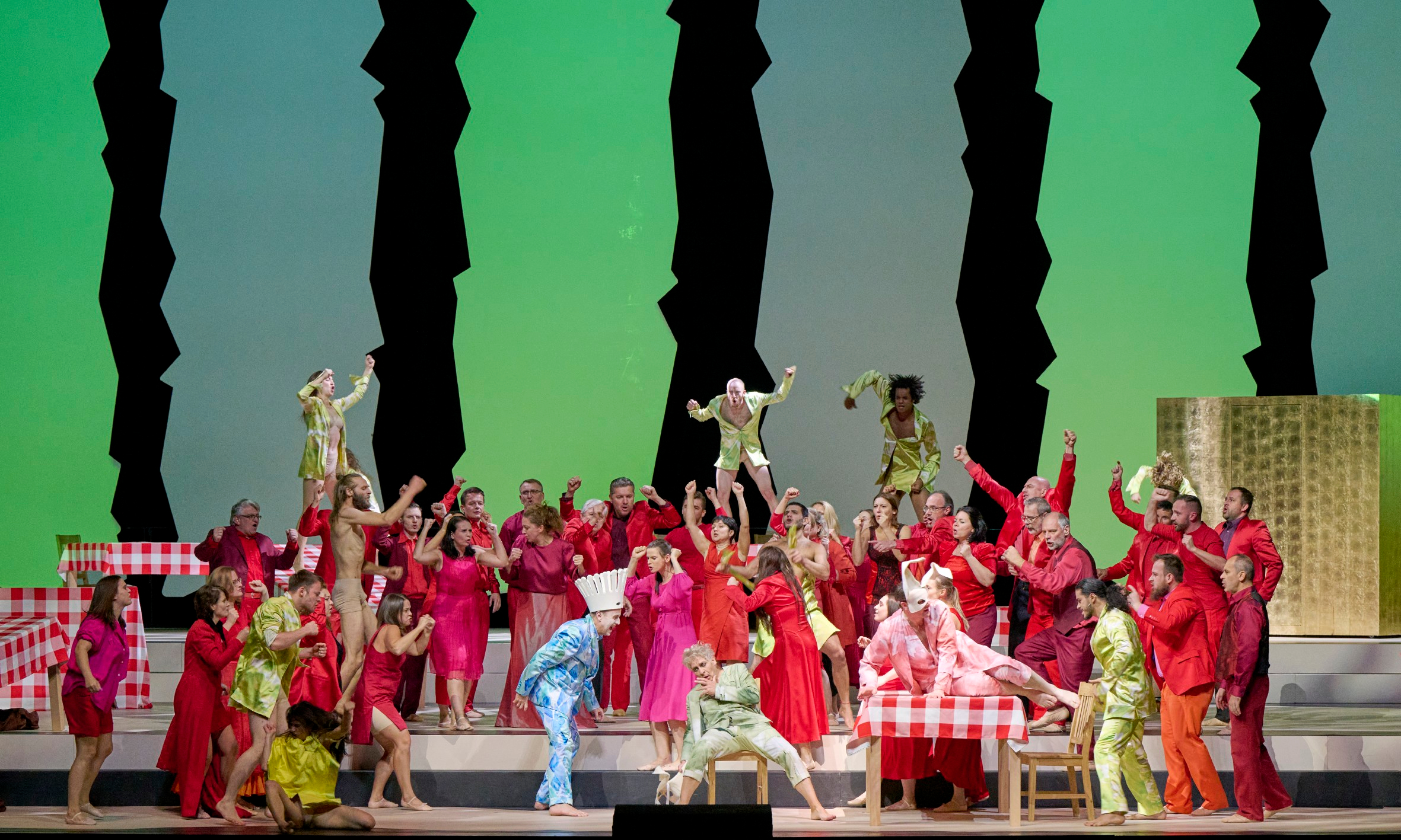 Eine große Gruppe bunt gekleideter Darsteller*innen tanzt ausgelassen auf der Bühne vor grünem Hintergrund mit schwarzen Silhouetten.