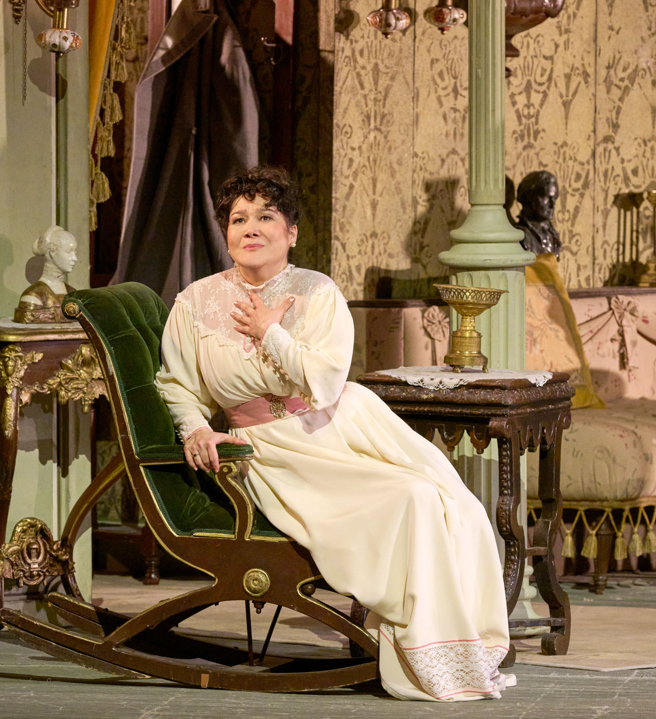 Eine Frau in hellem Kleid sitzt auf einem grünen Sessel in einem reich dekorierten Raum und blickt verträumt mit der Hand auf der Brust.