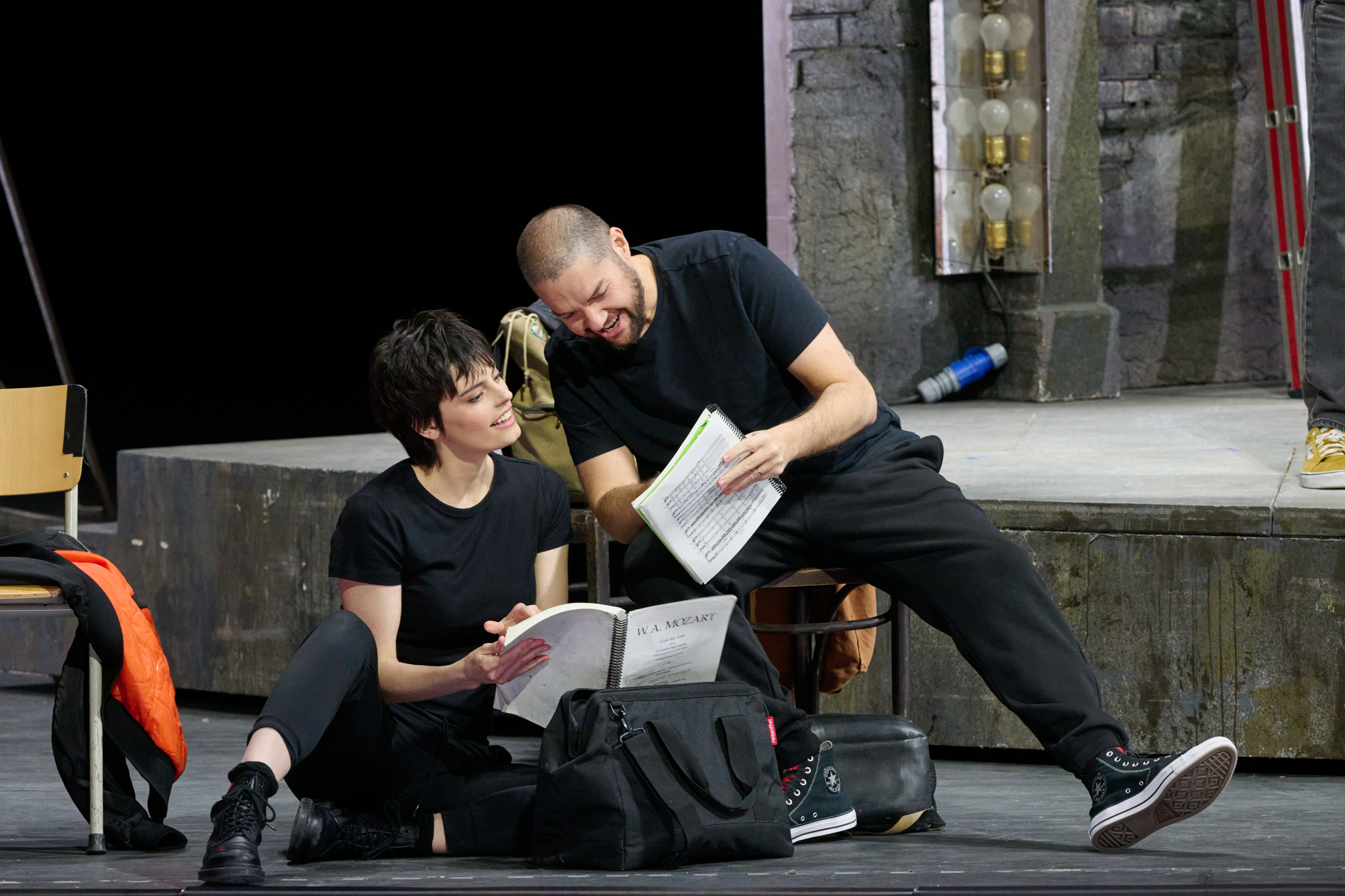 Zwei Personen in schwarzer Kleidung sitzen auf dem Bühnenboden und blättern gemeinsam durch Notenhefte oder Skripte, vertieft und lachend bei der Probe.