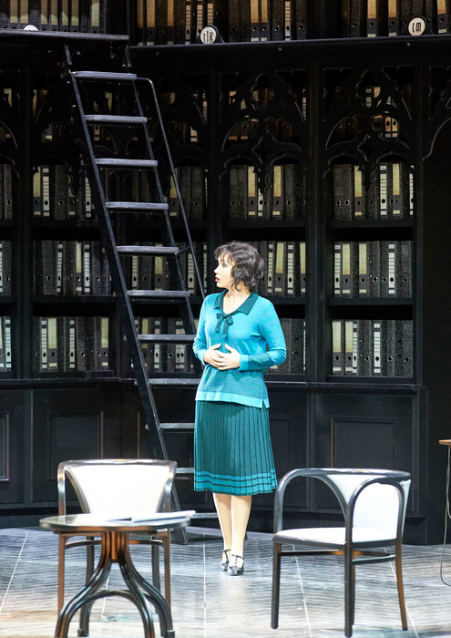 Eine Frau im türkiser Kleidung steht zwischen zwei Sesseln und vor einer dunklen Bücherwand.