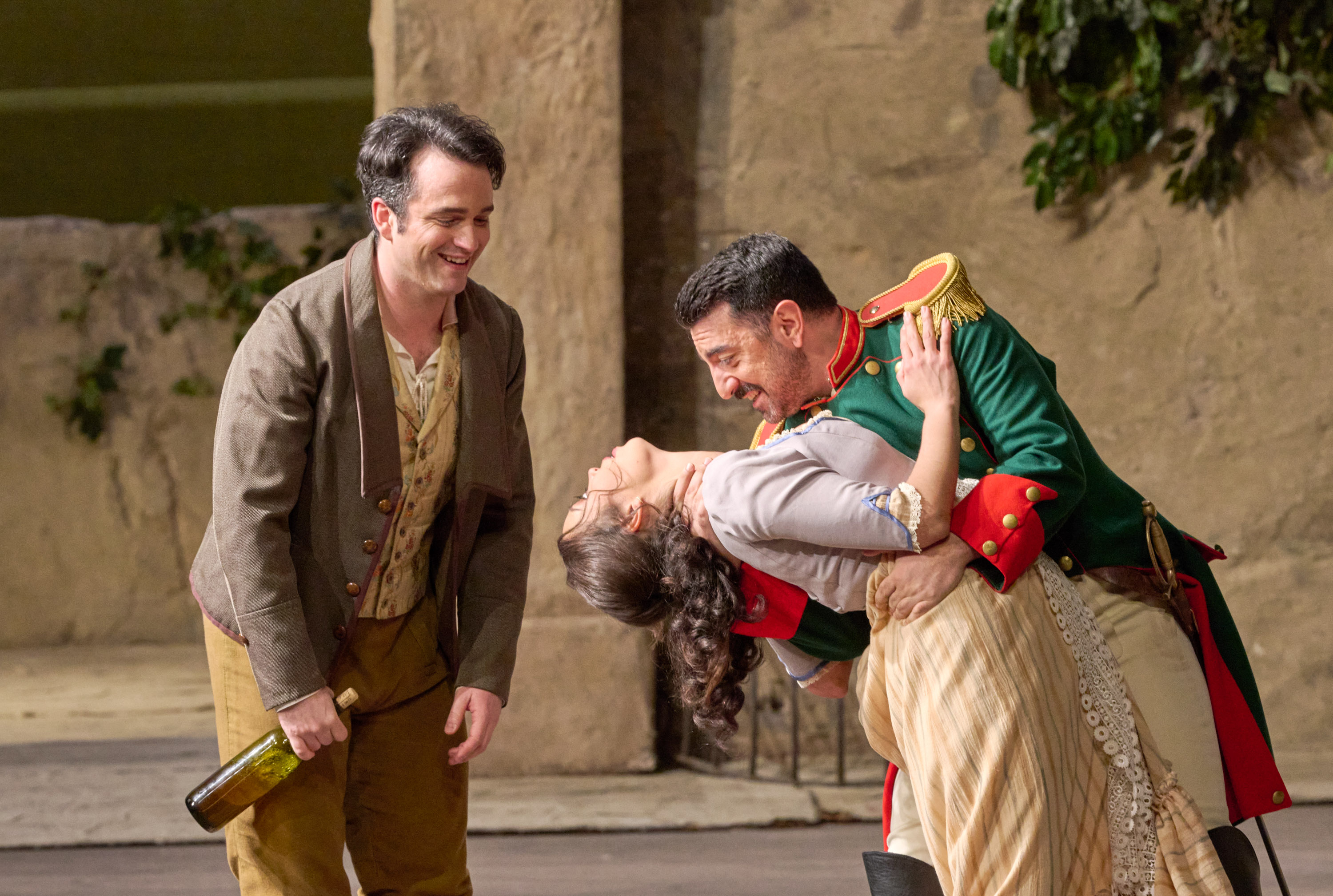 Ein Soldat taucht eine Frau in einer verspielten Tanzpose, während ein lachender Mann mit Flasche danebensteht.