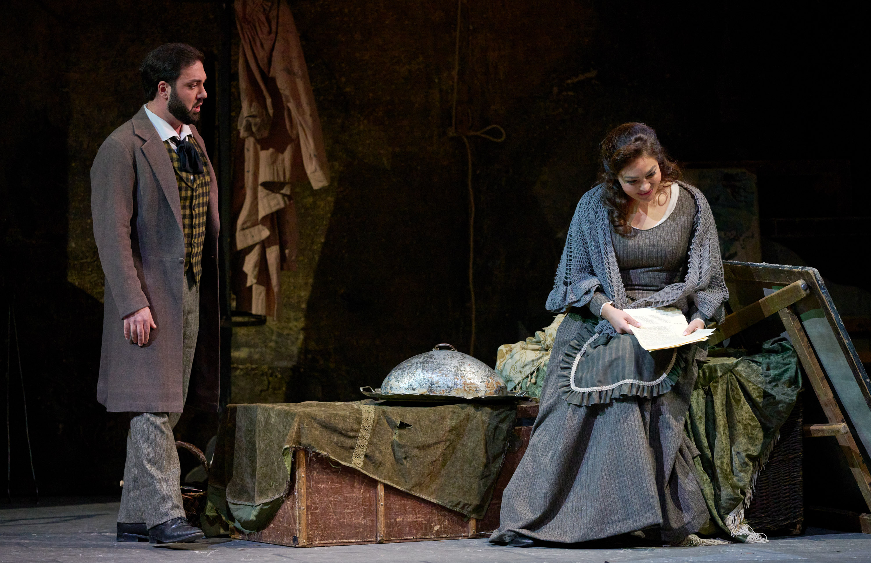 Eine Frau in grauer Kleidung sitzt und liest einen Brief, während ein Mann im Mantel sie mit ernster Miene anschaut.