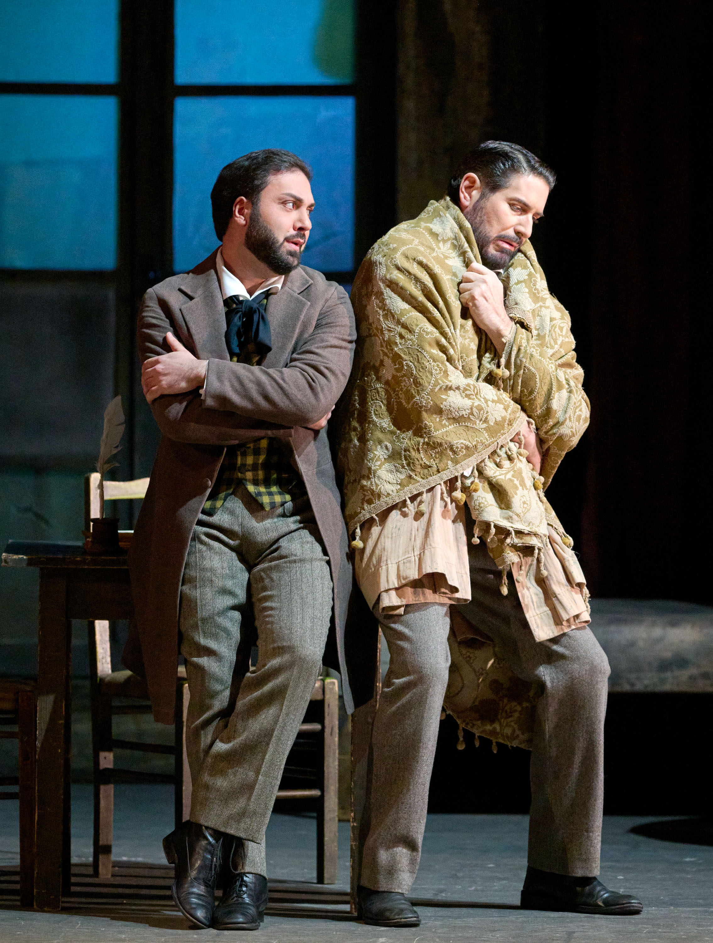 Zwei Männer in historischer Kleidung stehen Rücken an Rücken; einer ist in eine Decke gehüllt, der andere wirkt nachdenklich.