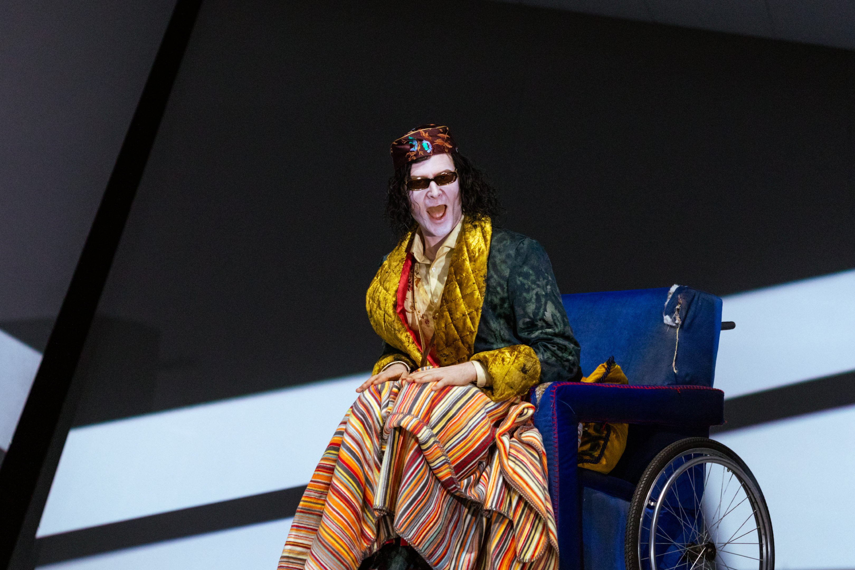 Eine Person in opulentem, bunt gemustertem Kostüm sitzt in einem Rollstuhl und ruft mit offenem Mund, Sonnenbrille und Kopfschmuck.