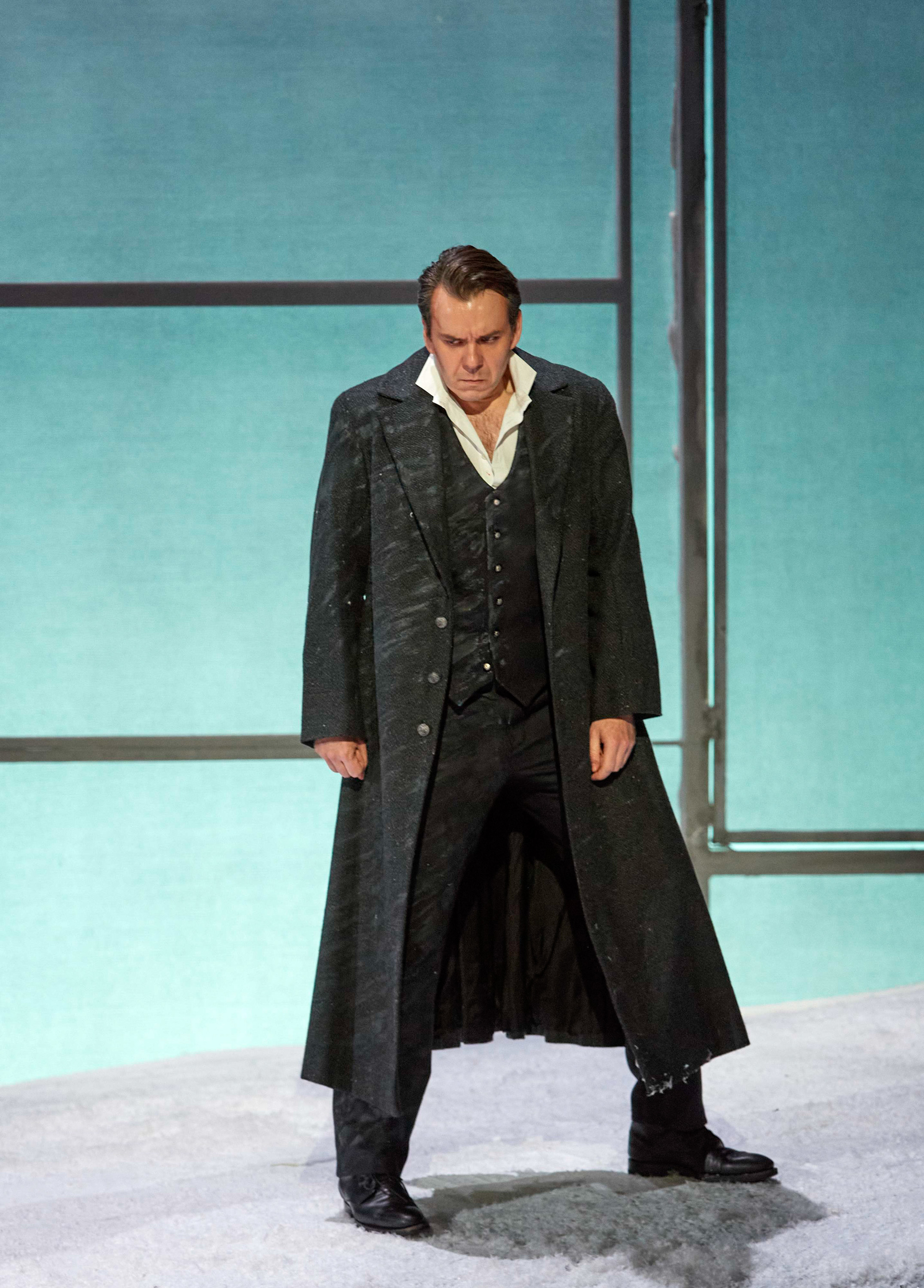 Ein Mann in einem langen, dunklen Mantel steht vor einer stilisierten, kühlen Bühnenkulisse und wirkt entschlossen oder angespannt.