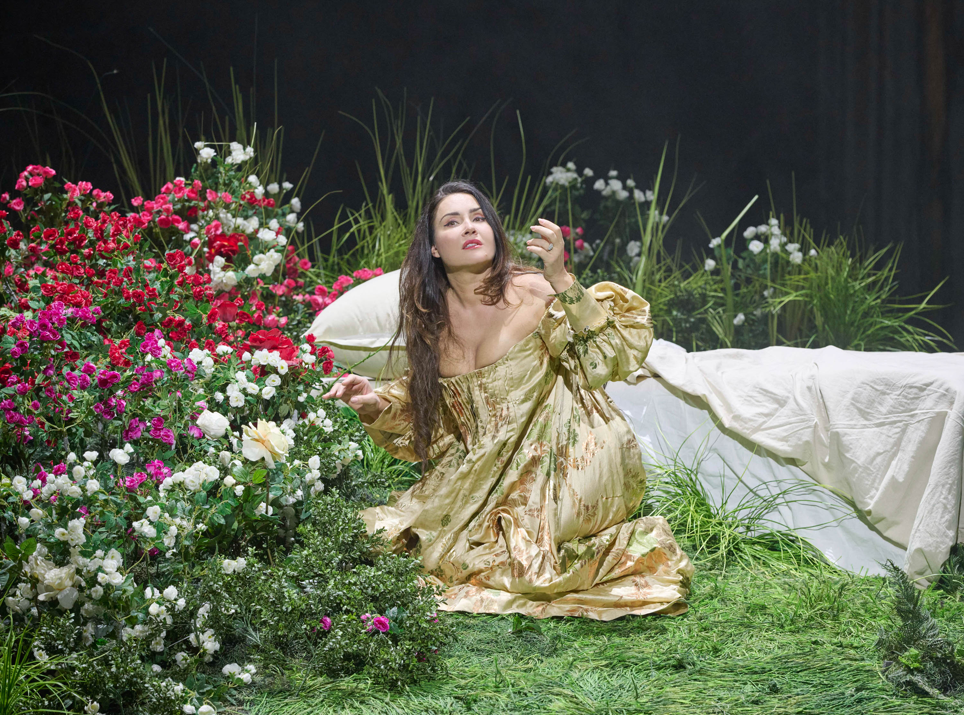 Eine Frau im goldenen Kleid kniet auf einer blumengeschmückten Wiese und blickt verträumt zu einer Rose in ihrer Hand.