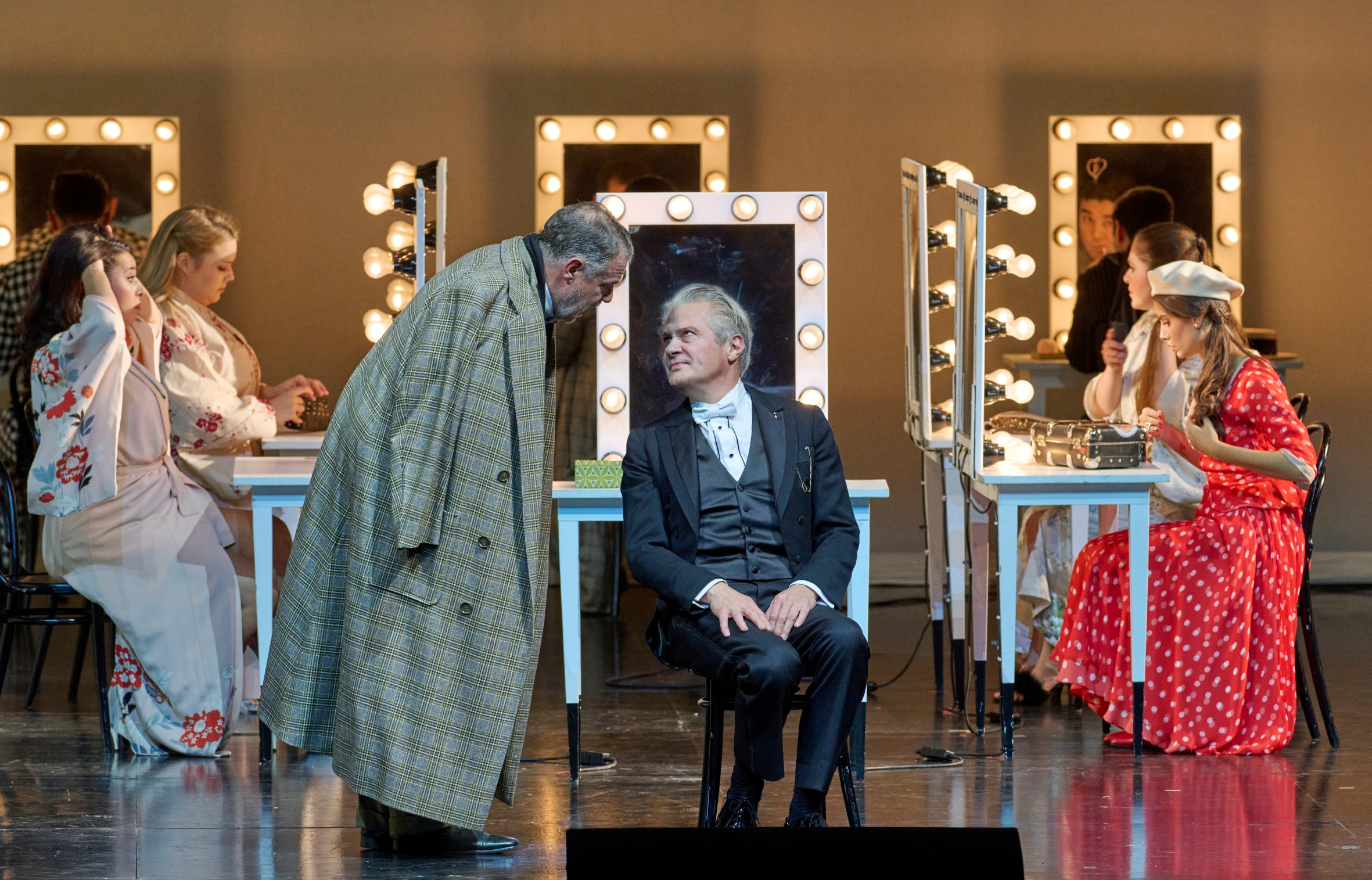 Ein Mann im karierten Mantel beugt sich zu einem elegant gekleideten Herrn, der vor einem beleuchteten Schminktisch sitzt; im Hintergrund sind weitere Personen in Vorbereitung.