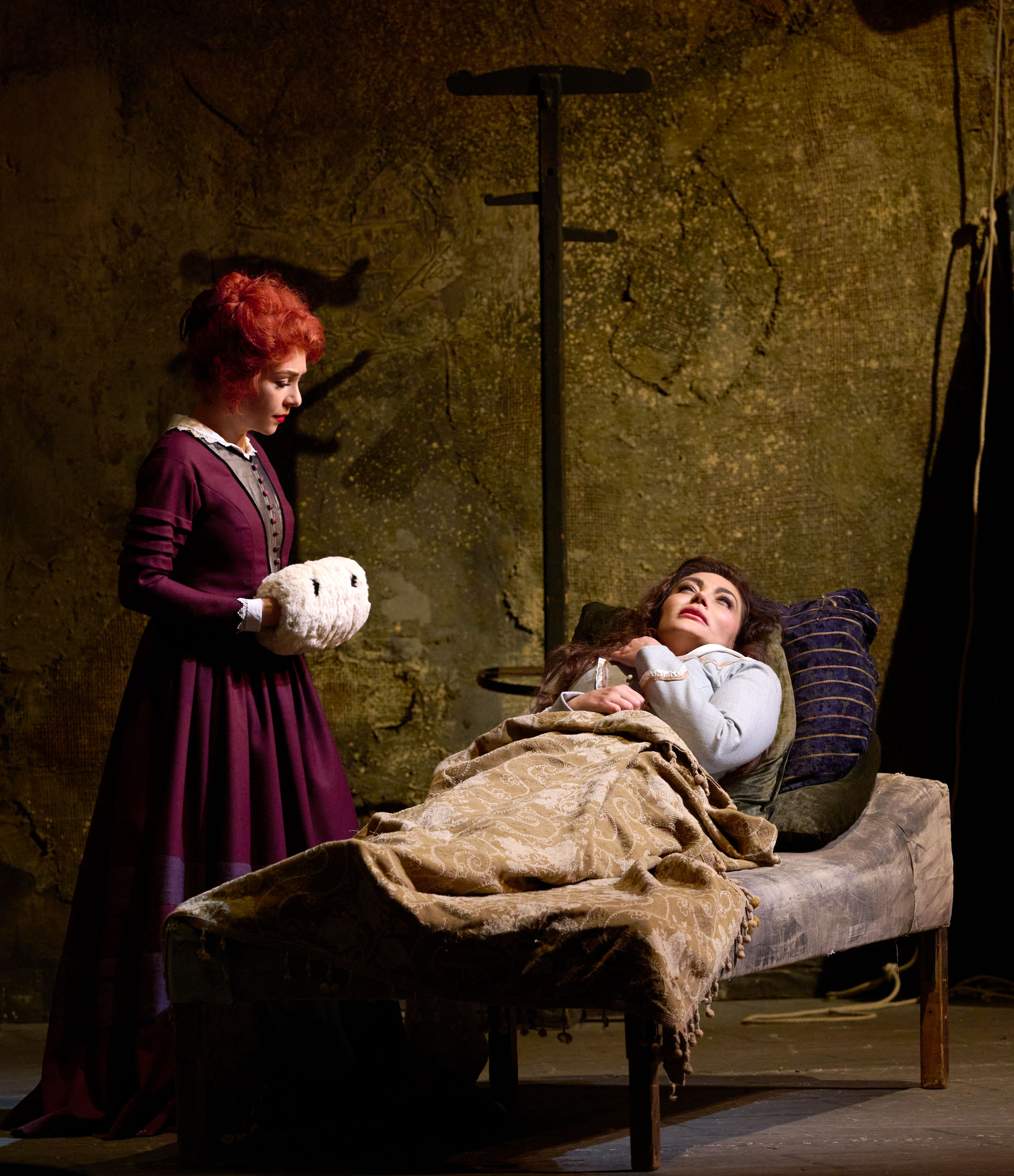 Eine Frau im Bett schaut erschöpft, während eine andere Frau in lila Kleid mit weißem Wärmekissen besorgt an ihrer Seite steht.