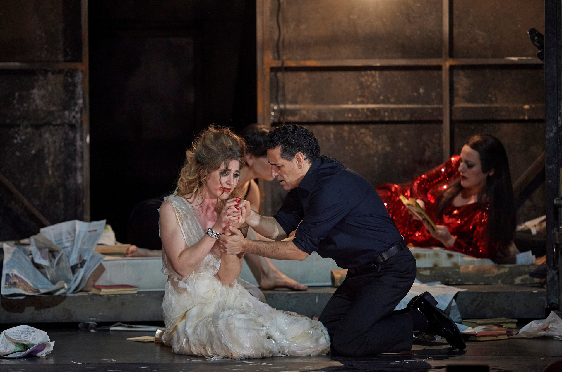 Eine Frau im weißen Kleid sitzt erschöpft und blutverschmiert am Boden, während ein Mann sich sorgenvoll zu ihr kniet und ihre Hand hält.