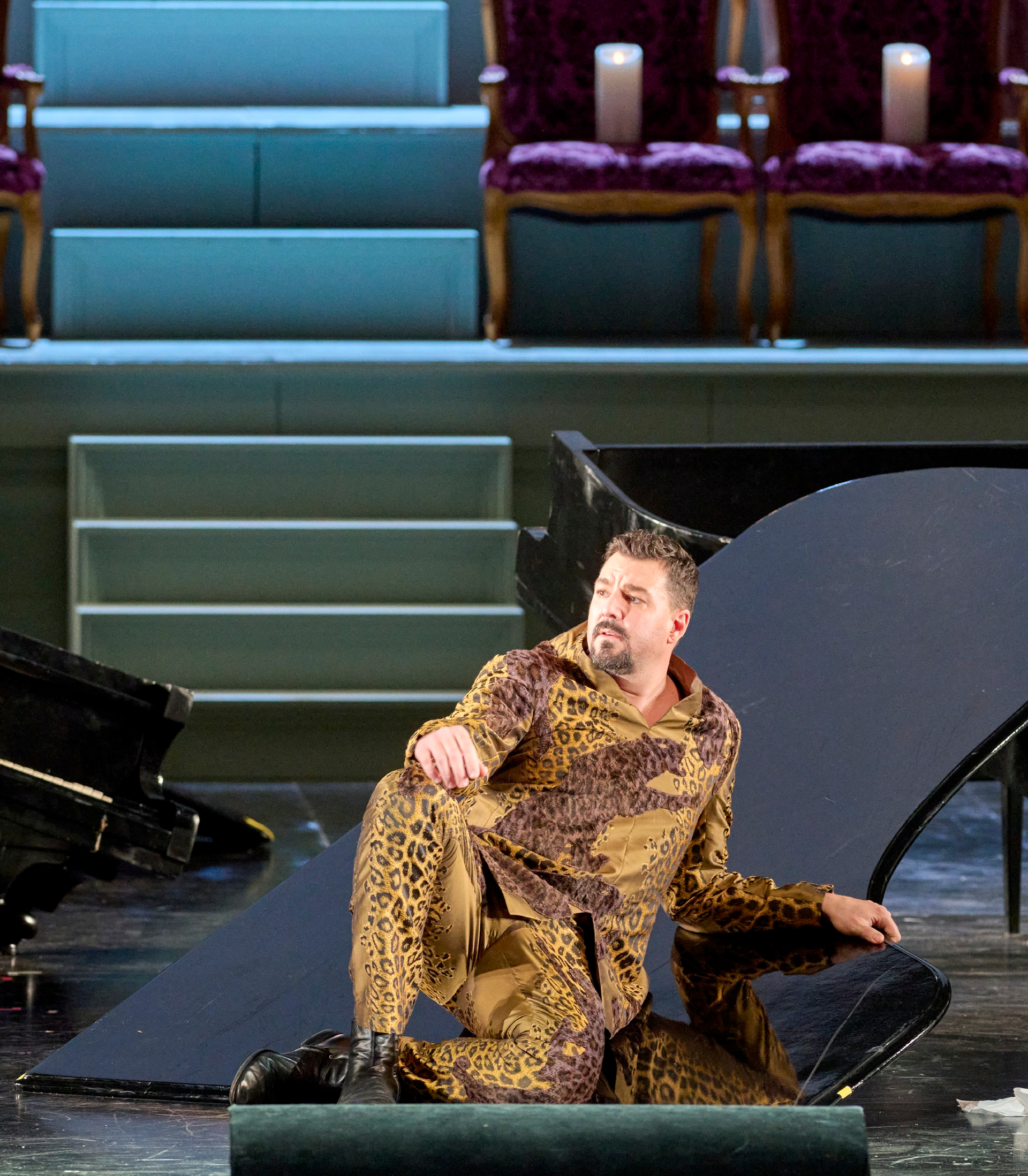 Ein Mann in gemustertem goldbraunen Anzug sitzt auf dem Boden vor einem umgestürzten Flügel und blickt nachdenklich zur Seite; im Hintergrund leere Theatersessel.