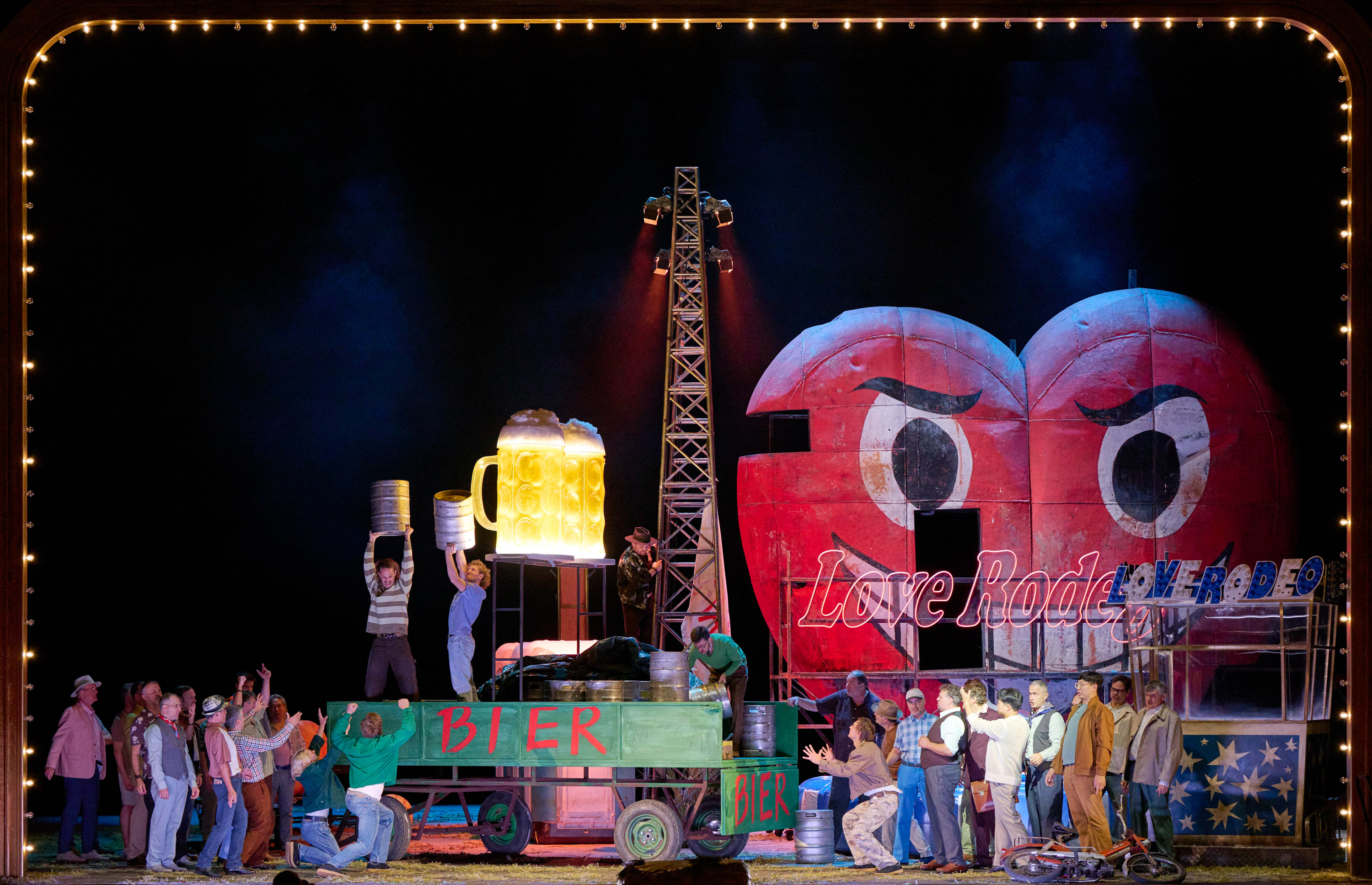 Auf einem Anhänger mit der Aufschrift "Bier" stehen zwei Männer mit riesigen Biergläsern. Neben dem Wagen stehen viele Menschen, die zu den beiden Männern hinaufblicken. Im Hintergrund sieht man die Stände einer Kirmes.