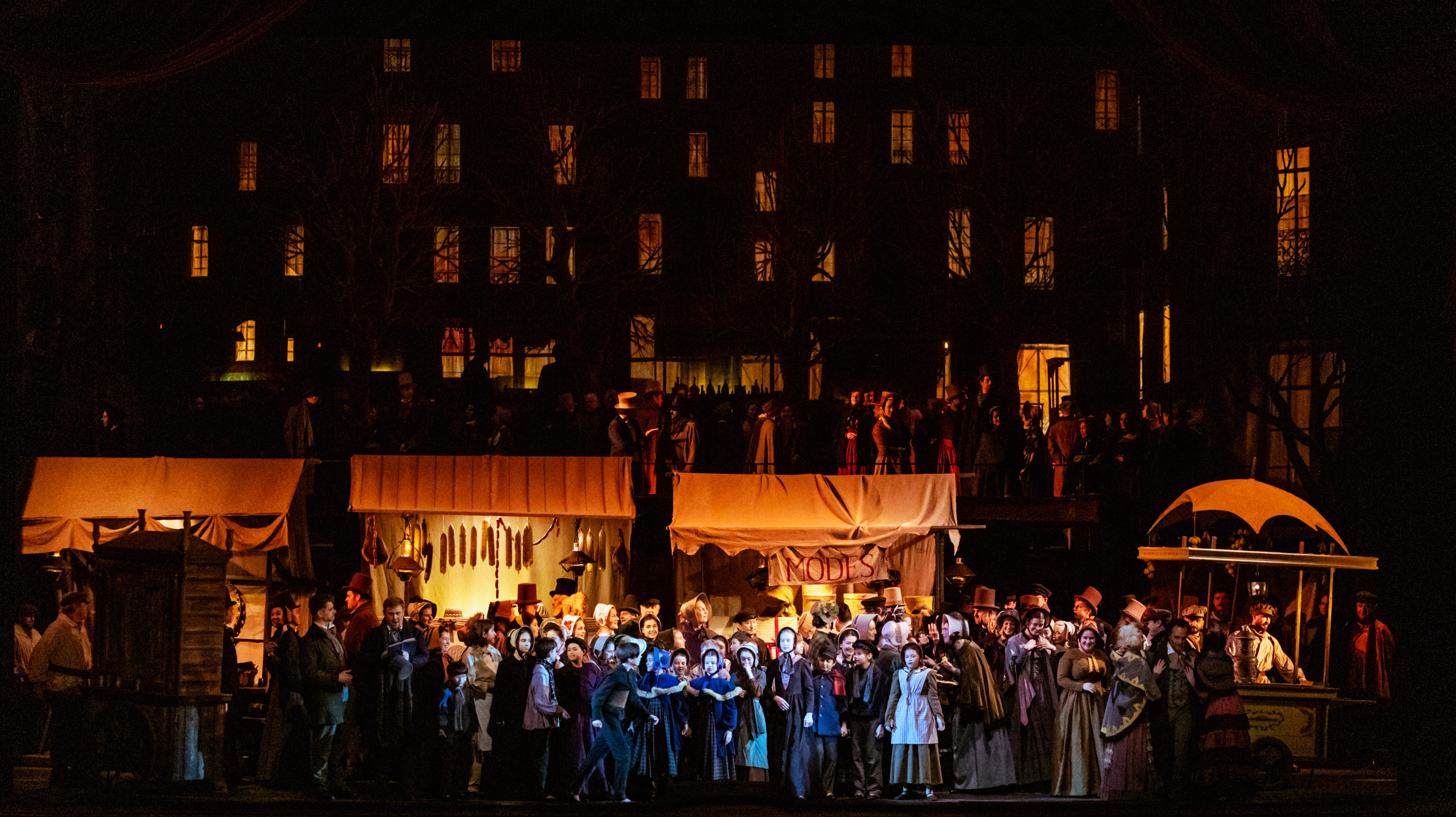 Eine große Menschenmenge in historischer Kleidung versammelt sich vor beleuchteten Fenstern und Marktständen in nächtlicher Szenerie.