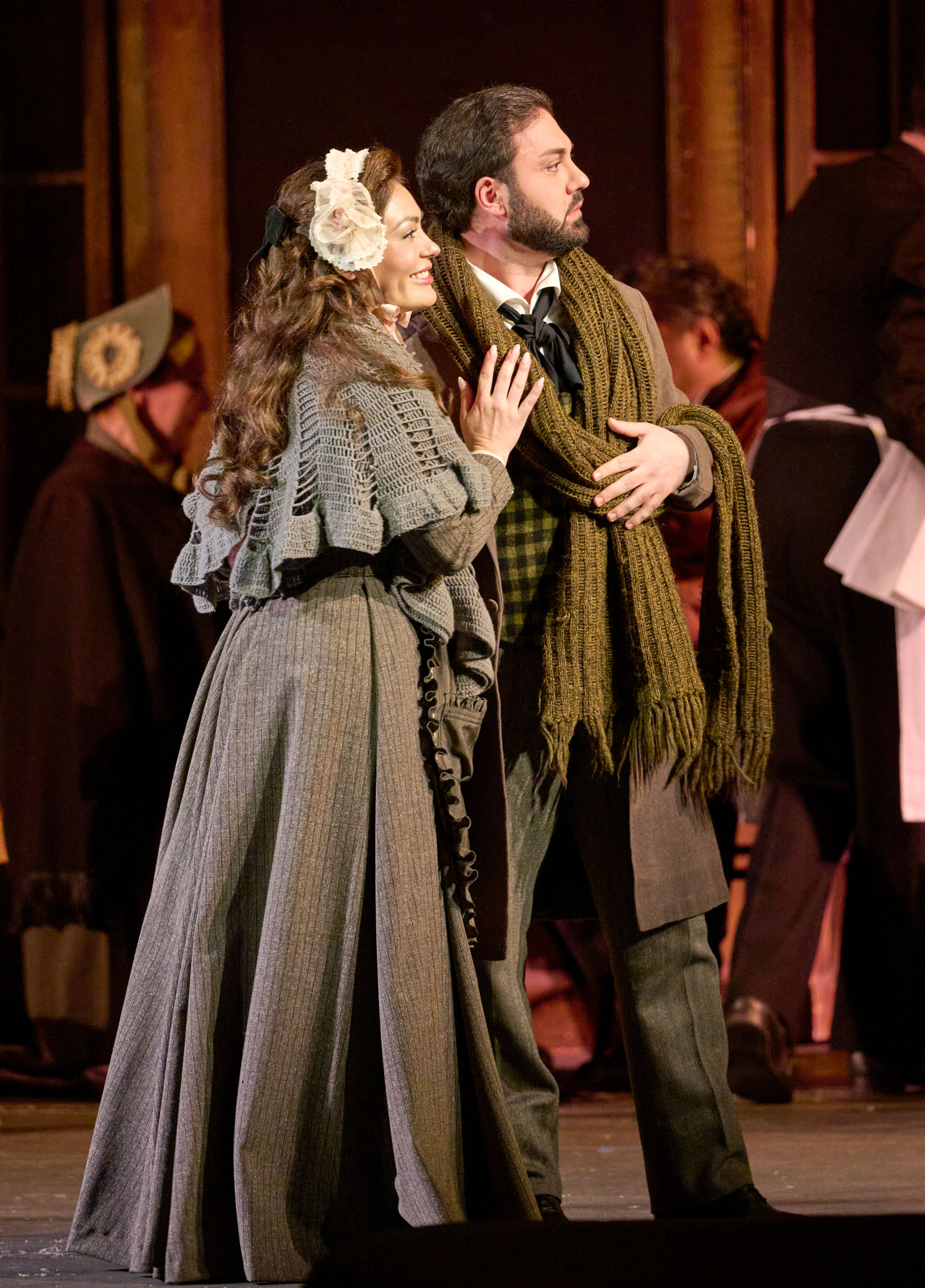 Ein Paar in historischer Kleidung steht eng beieinander und blickt erwartungsvoll in dieselbe Richtung, umgeben von weiteren Personen.