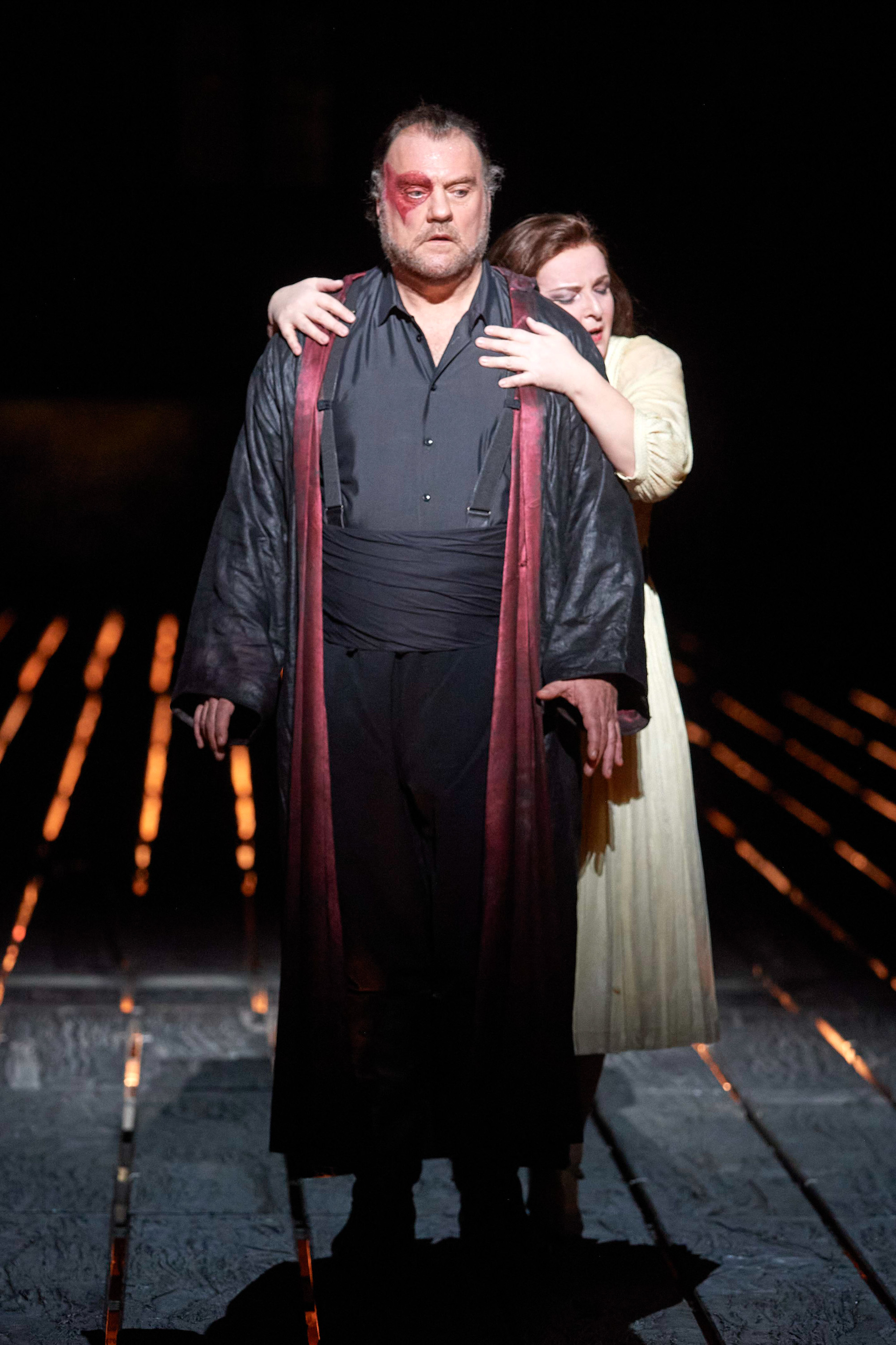 Eine Frau schmiegt sich von hinten an einen dunkel gekleideten Mann mit einer Gesichtsverletzung.