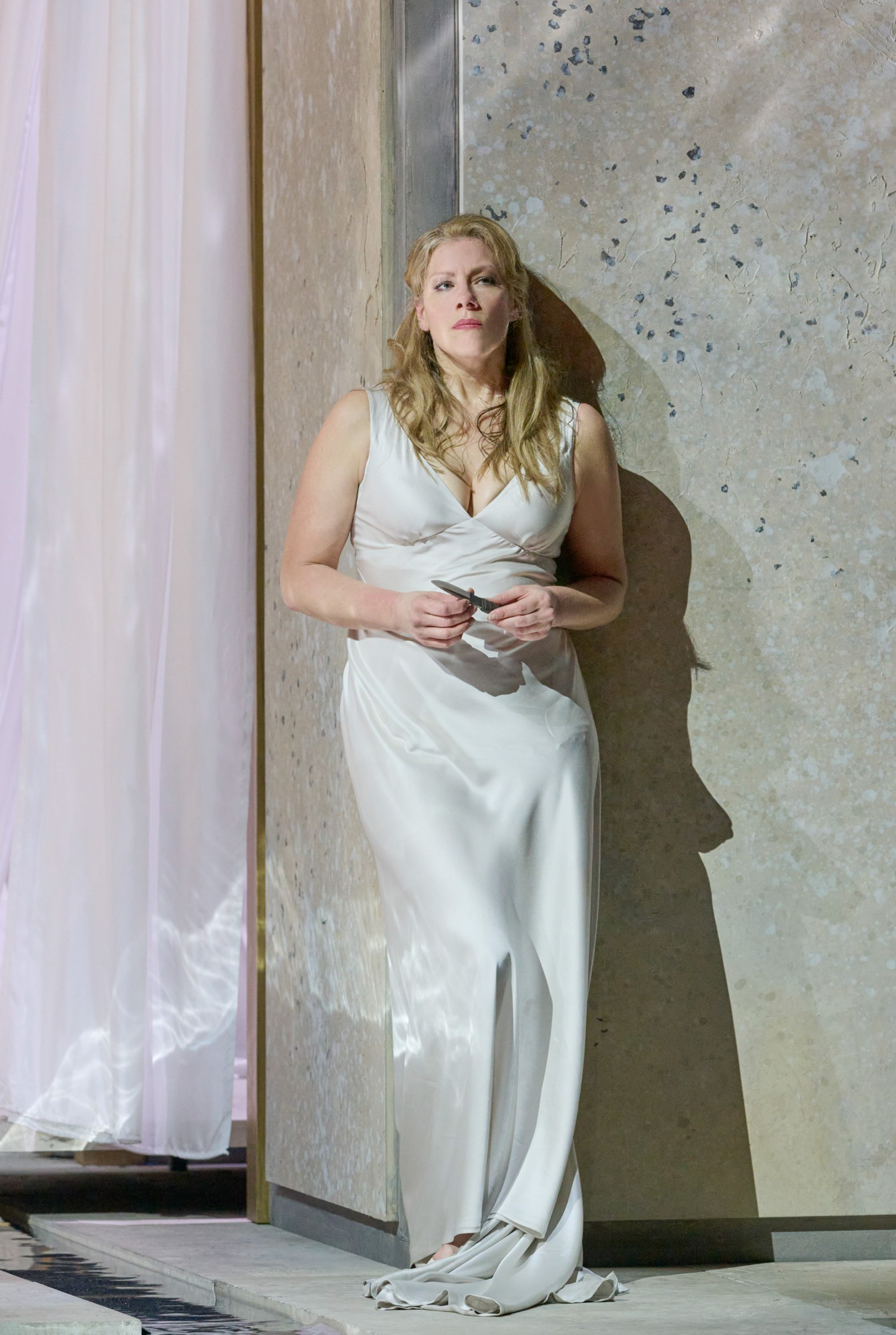 Eine Frau in einem weißen Kleid lehnt nachdenklich an einer hellen Wand mit schimmernden Reflexen.