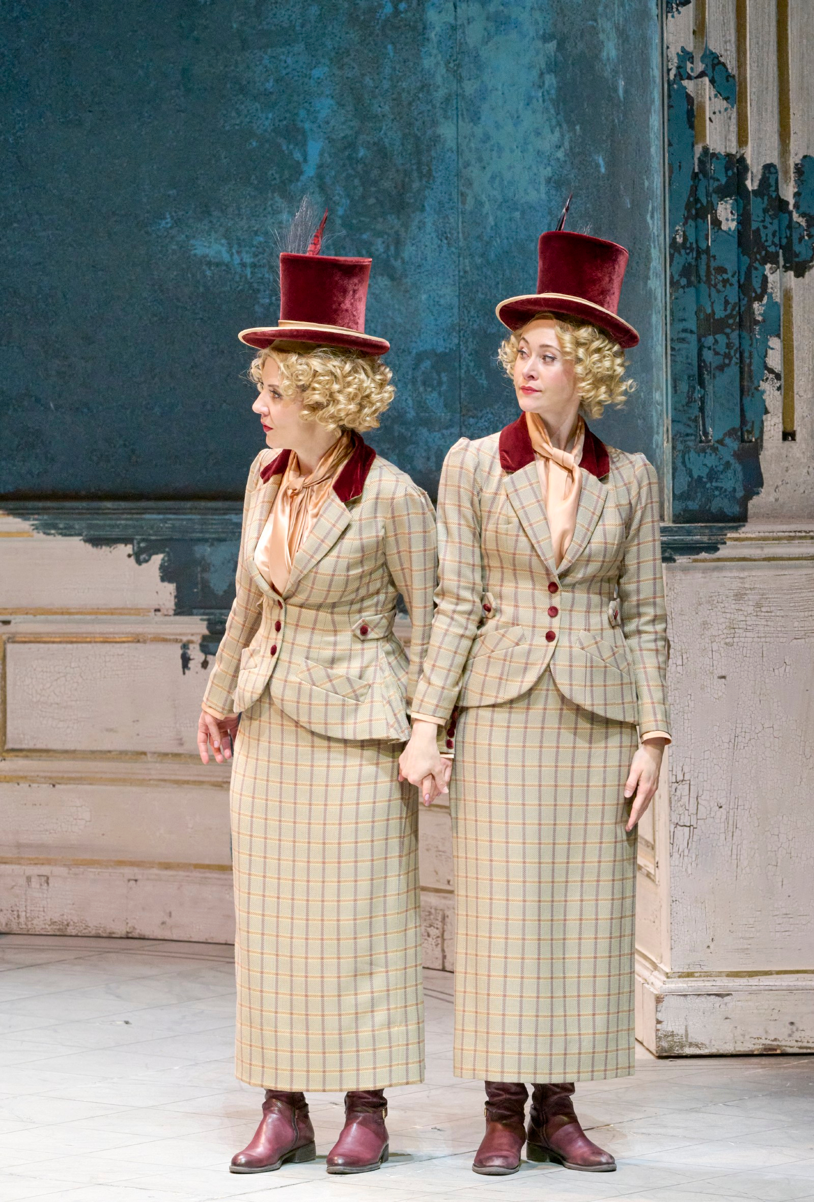 Zwei identisch gekleidete Frauen mit Lockenperücken und roten Zylindern stehen Hand in Hand vor einer blauen, abgeblätterten Wand.