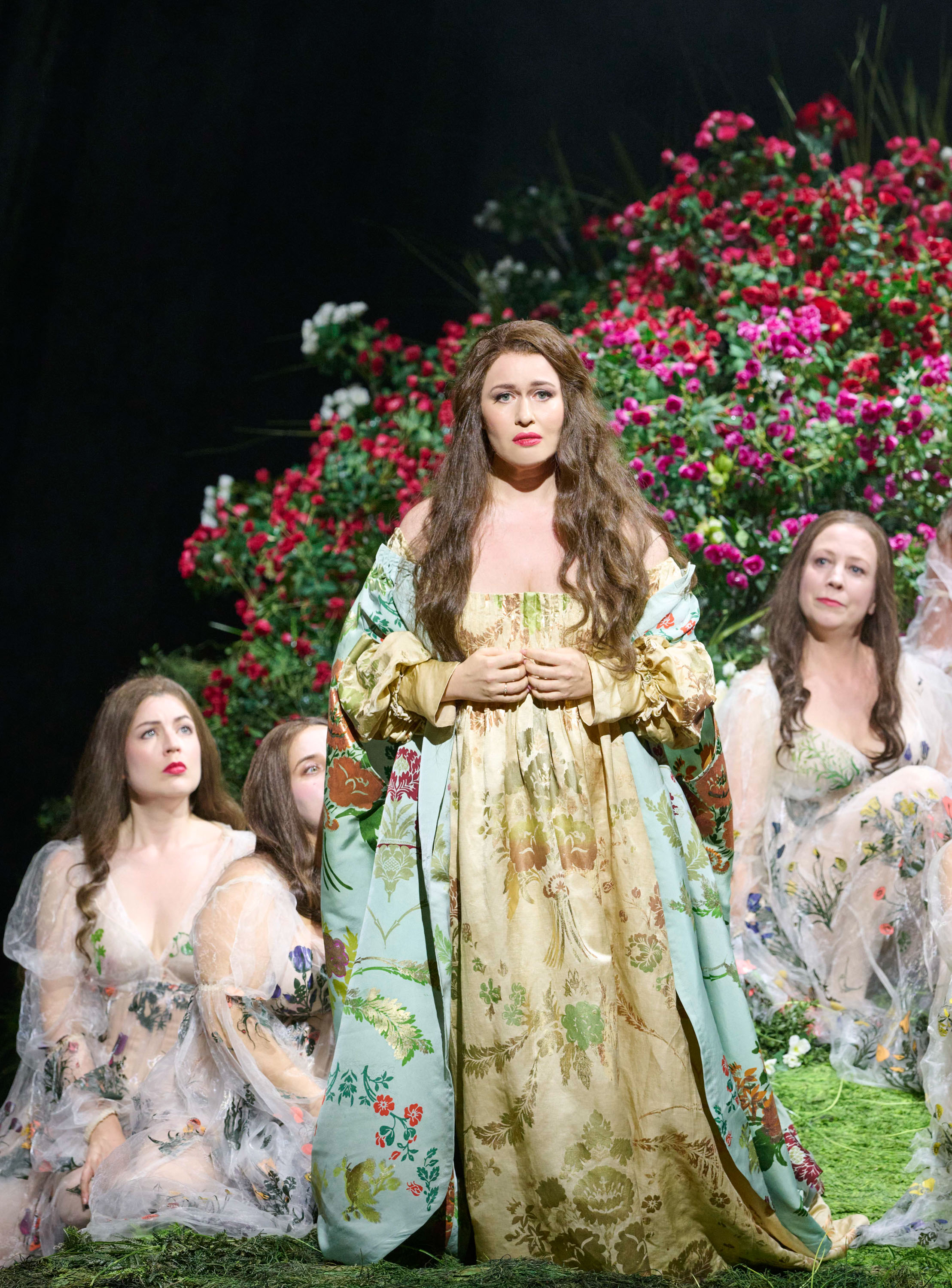 Eine langhaarige Frau steht in einem langen hellgelben Gewand in der Mitte. Um sie herum stehen weitere Frauen. Im Hintergrund sind viele Blumen.