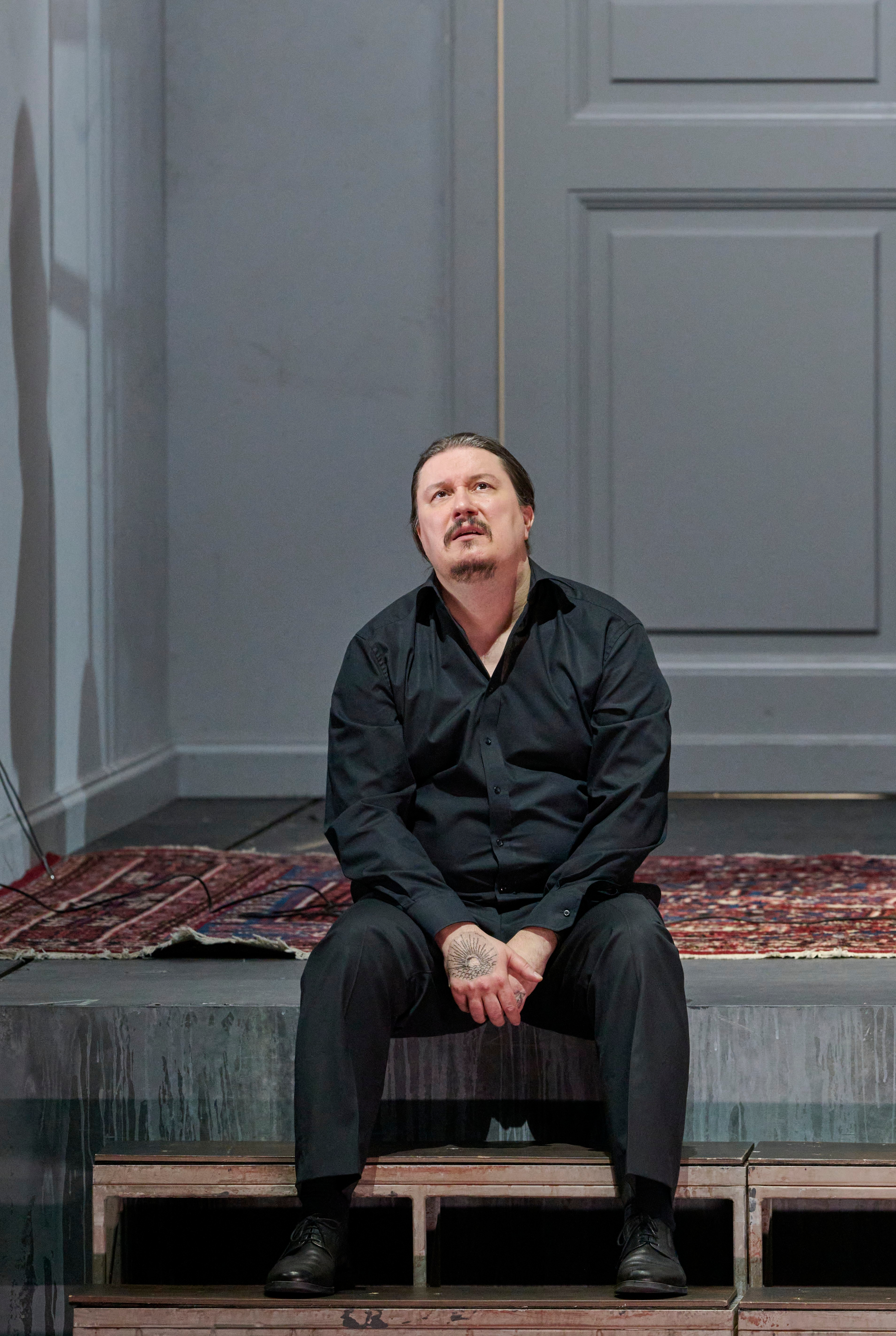 Ein Mann in schwarzer Kleidung sitzt allein und niedergeschlagen auf einem Teppich vor einer grauen Wand.