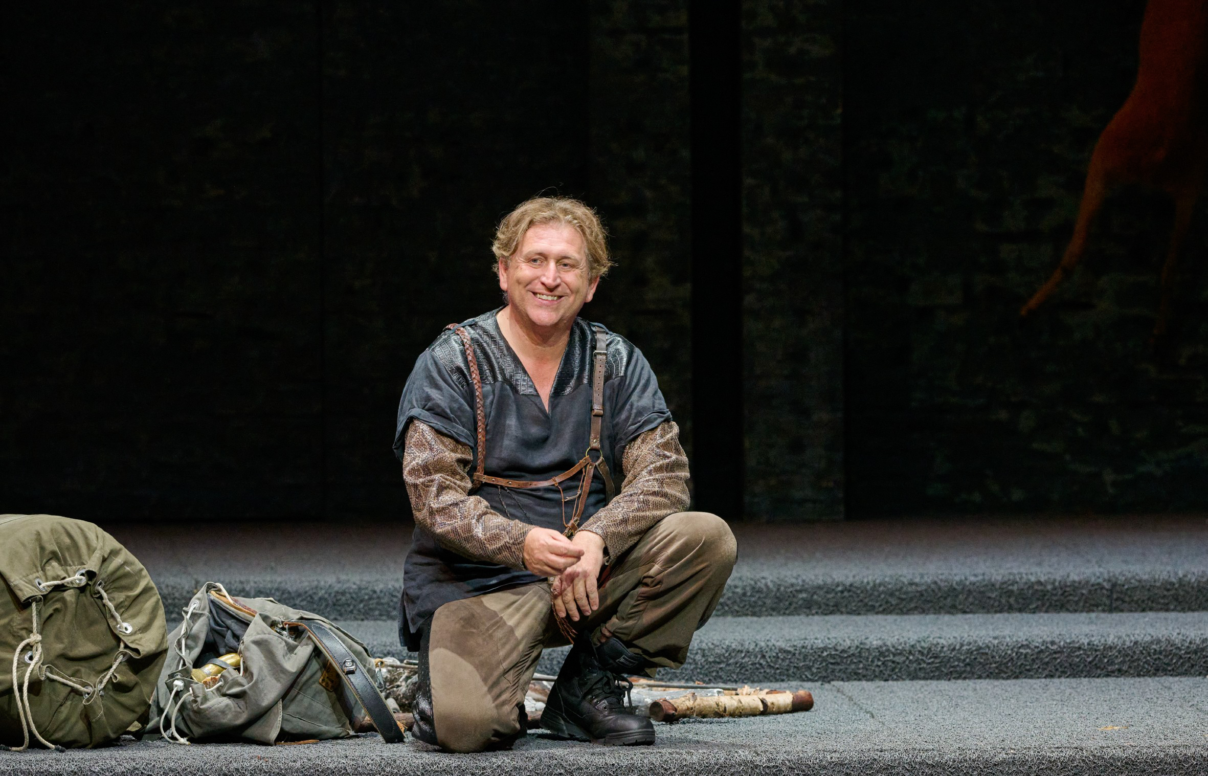 Ein Mann in dunkler Kleidung kniet auf einer leeren Bühne und blickt nach vorne.