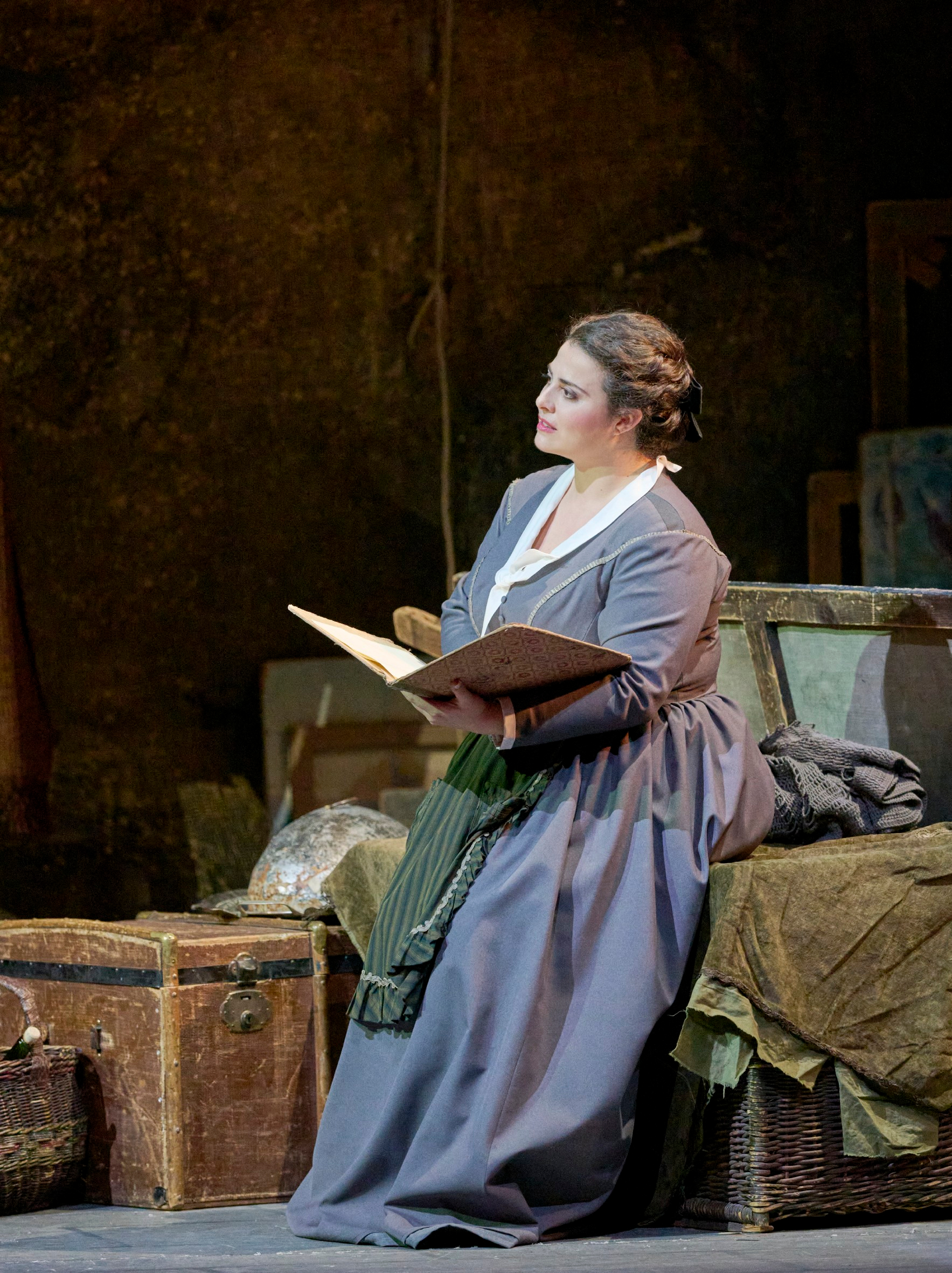 Eine Frau in historischer Kleidung sitzt in einem spärlich möblierten Raum und hält ein geöffnetes Buch in der Hand.