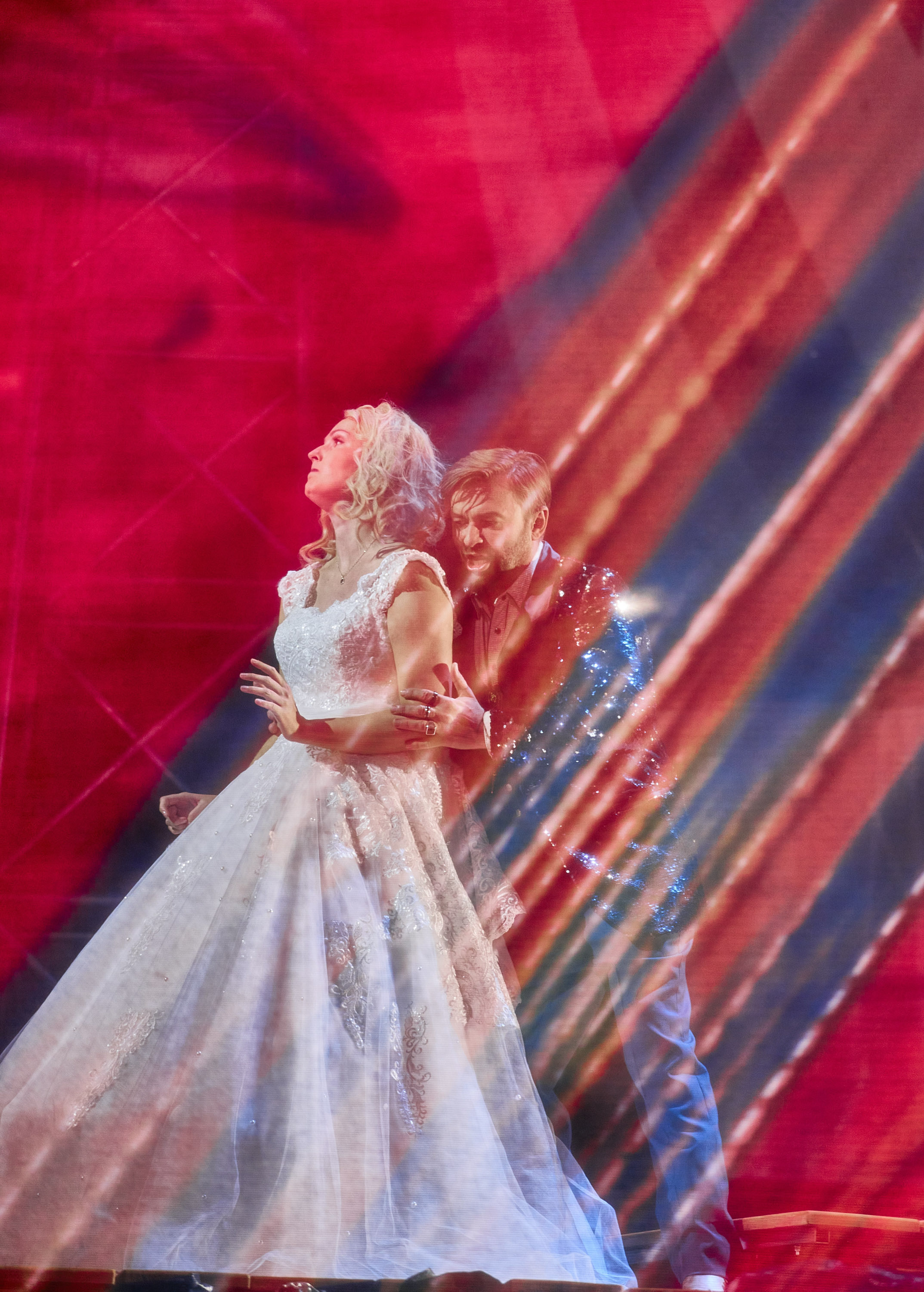 Eine Frau in einem prunkvollen weißen Kleid steht in einem rot und blau beleuchteten Raum. Hinter ihr steht ein Mann, der sich an ihr festhält.