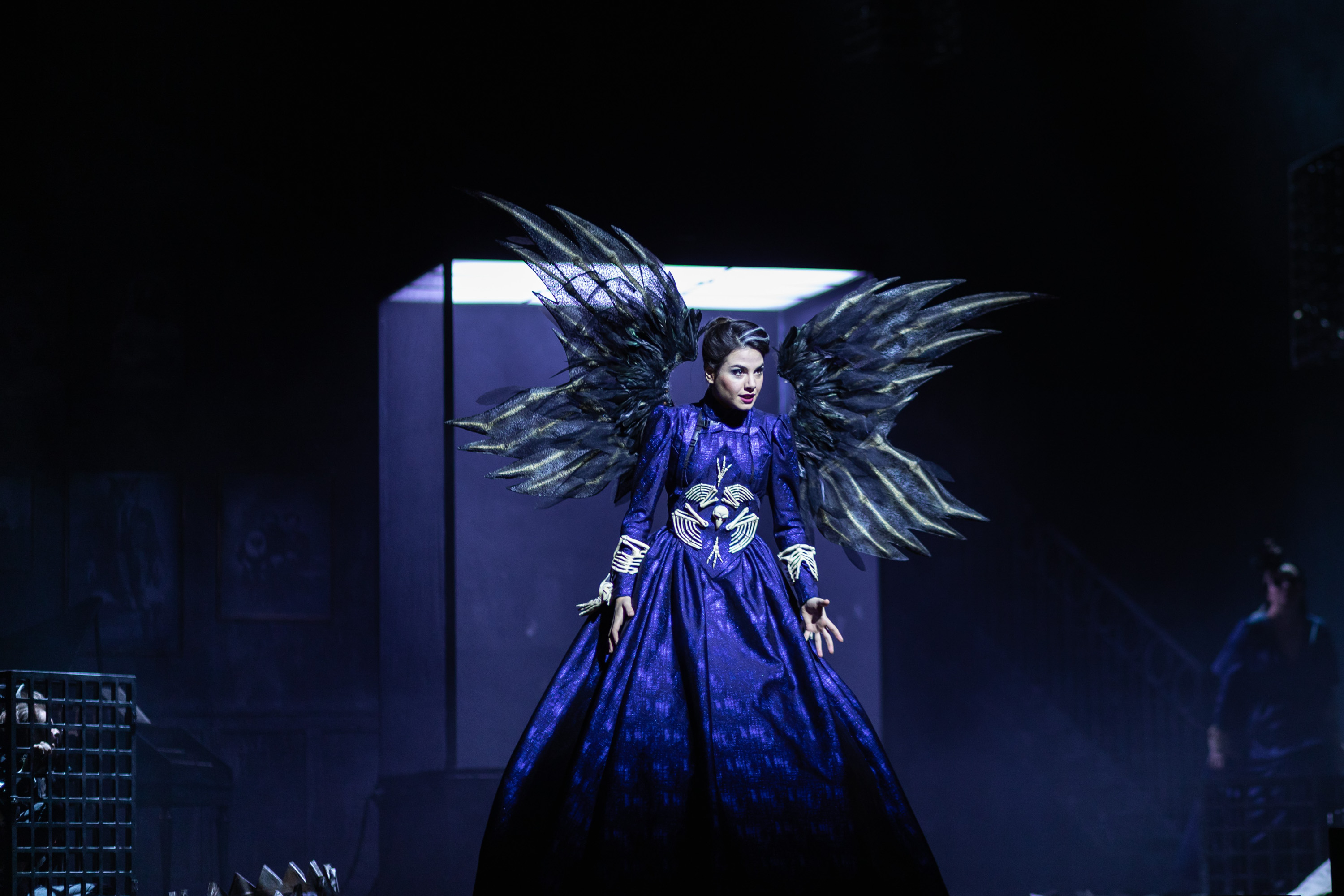 Eine Frau in einem blauen Kleid mit kunstvollen schwarzen Flügeln steht dramatisch auf der Bühne, die Arme weit ausgebreitet.