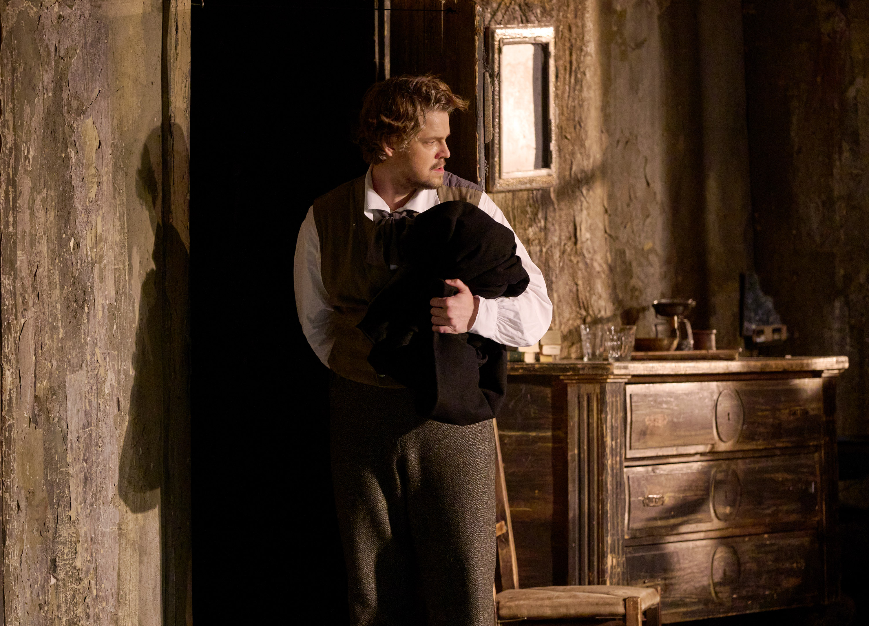 Ein Mann in Weste steht in einem düsteren Raum und blickt nachdenklich zur Seite, während er ein schwarzes Kleidungsstück im Arm hält.